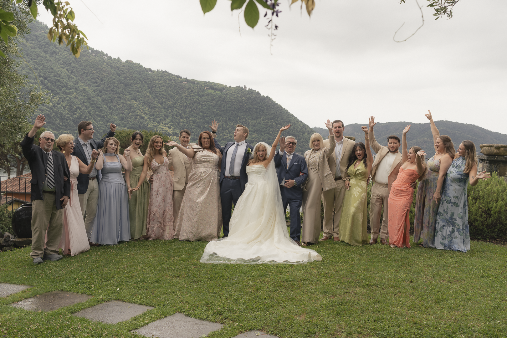 Gruppo di persone in abiti eleganti e da matrimonio che celebrano all'aperto, con scenografia naturale di colline verdi sullo sfondo.