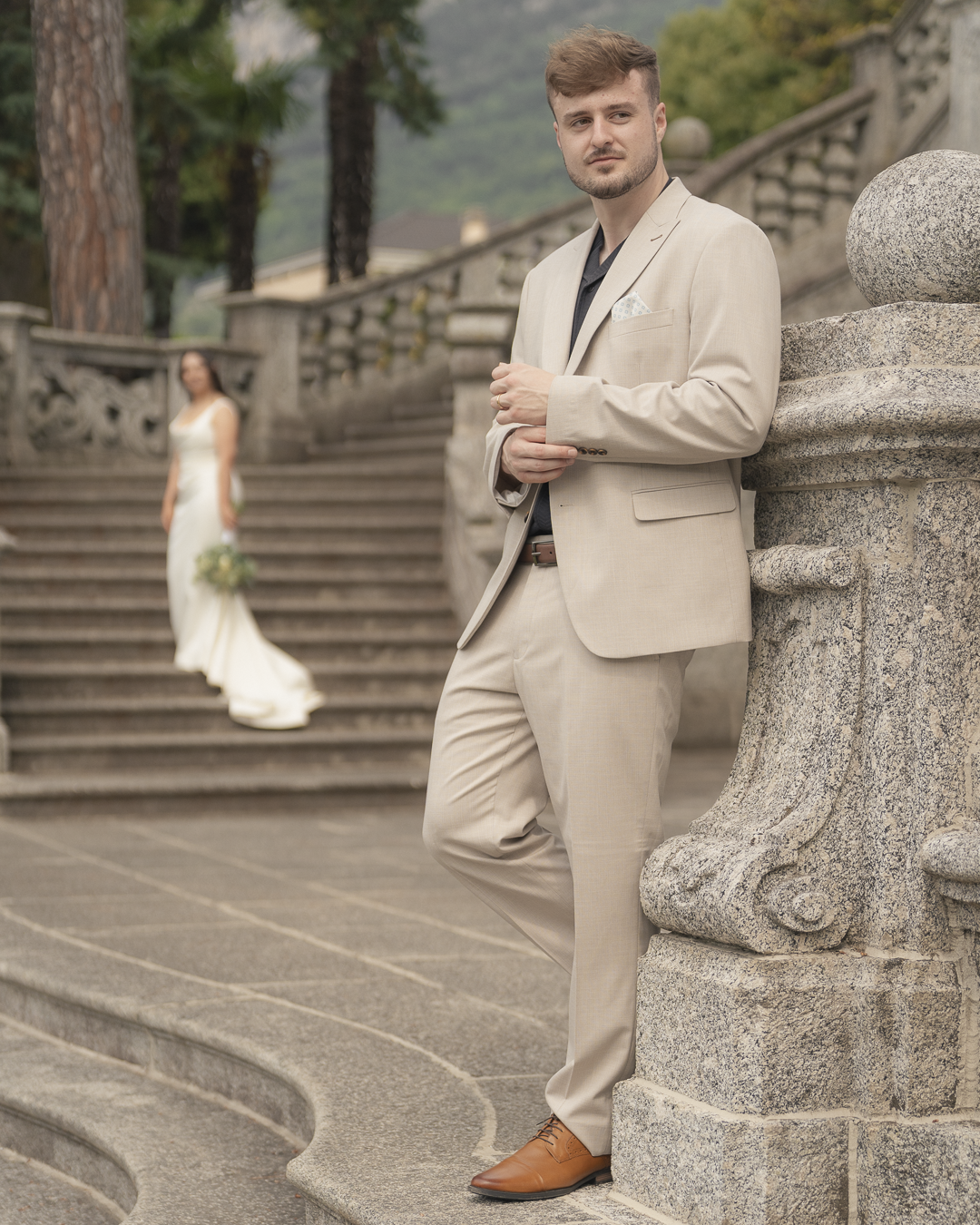 Un uomo in abiti eleganti beige si appoggia a una balaustra di pietra, con uno scenario di un cornicione d'epoca e un giardino sullo sfondo. Sullo sfondo si vede anche una donna in abito bianco con bouquet, probabilmente una sposa.