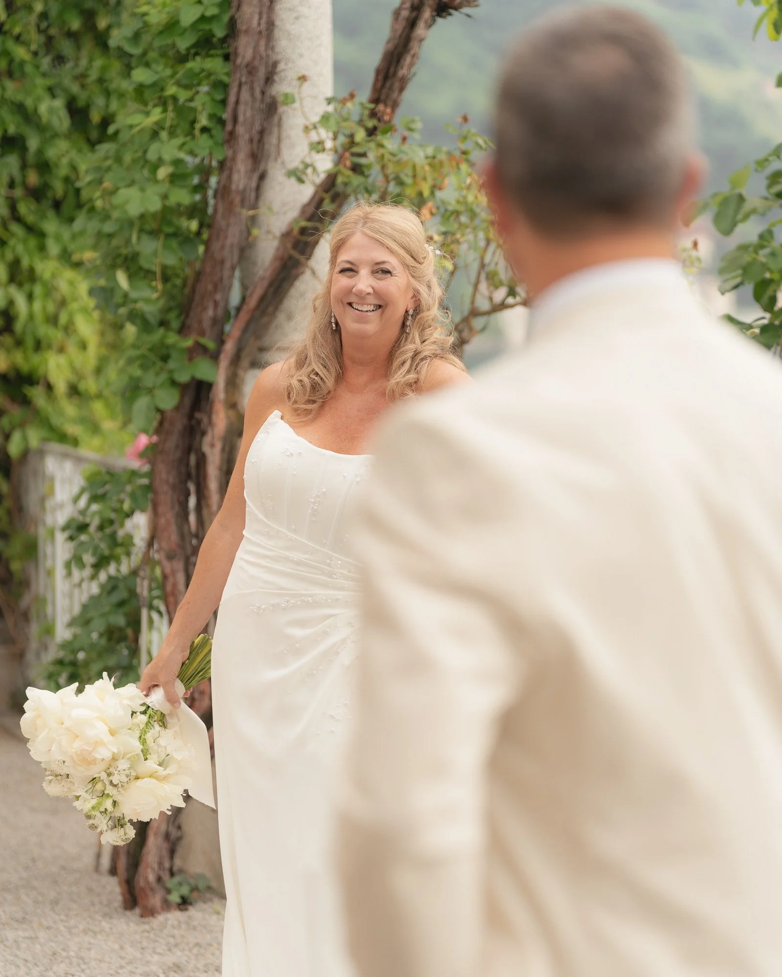 Una coppia di persone durante un matrimonio, con una donna sorridente che tiene un bouquet di fiori bianchi, in un ambiente all'aperto con alberi e natura.