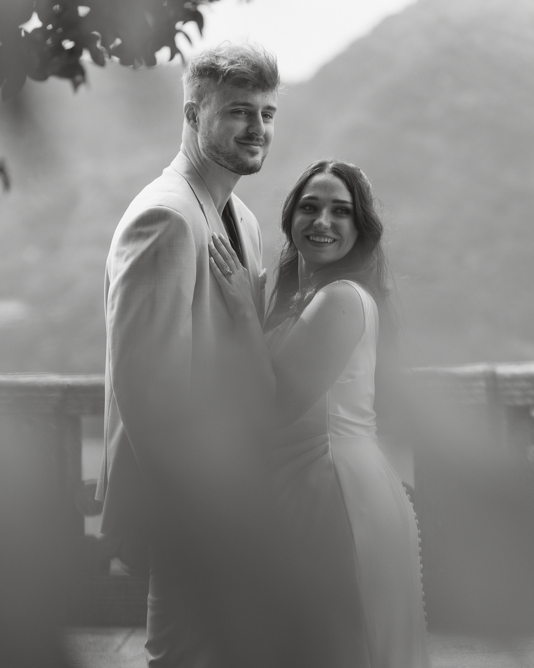 Una coppia sorridente, un uomo e una donna, si abbracciano e si tengono per mano all'aperto con uno sfondo di montagne e vegetazione, in un'immagine in bianco e nero.