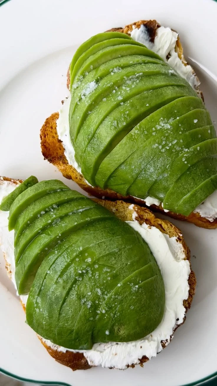 Two slices of toasted bread topped with creamy spread, sliced green avocado, and a sprinkle of salt.