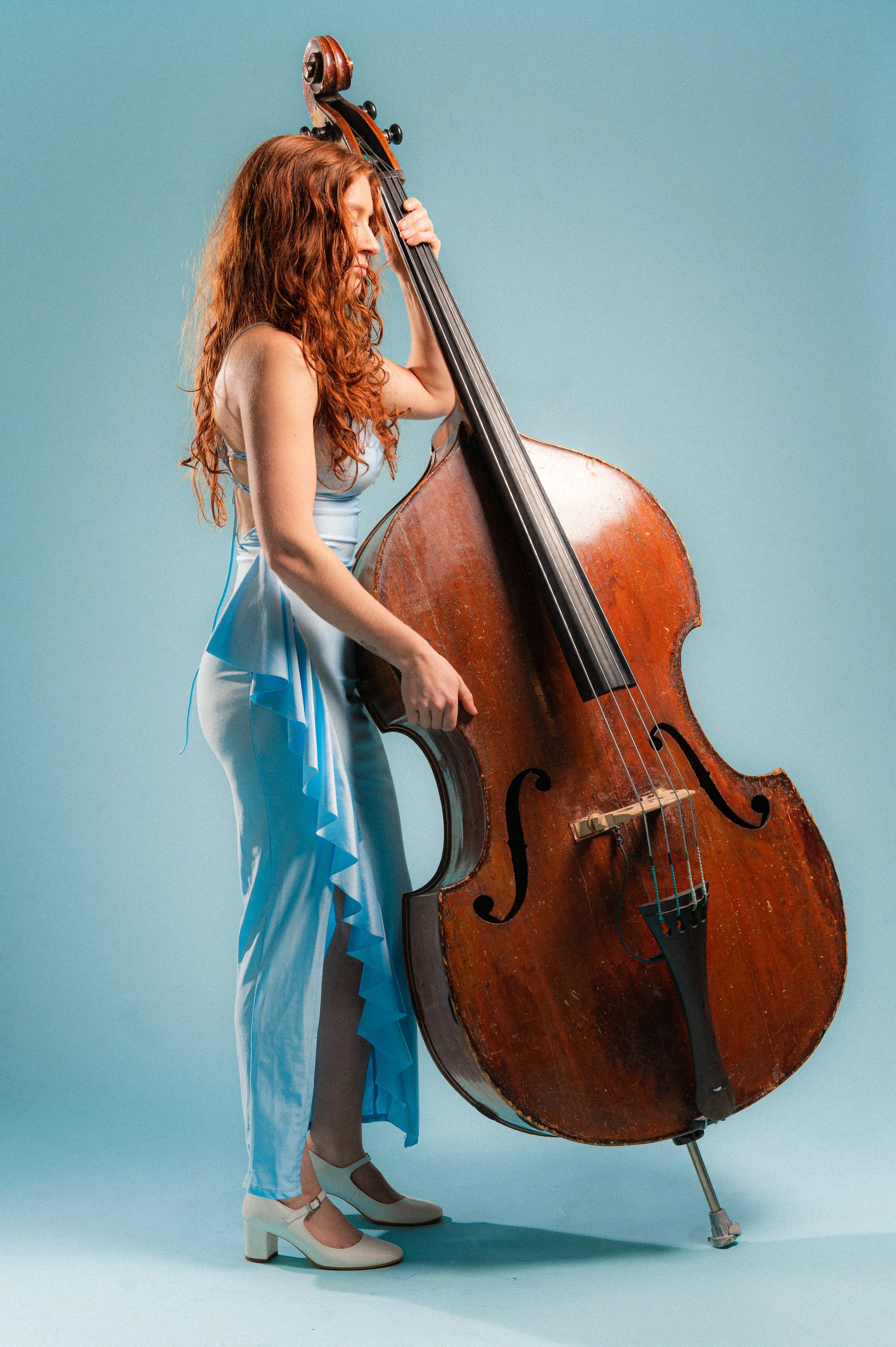 A woman with long, curly red hair playing a large upright double bass against a plain blue background. She is dressed in a light blue gown with ruffles and white heeled shoes, with her eyes closed and head tilted slightly downward.