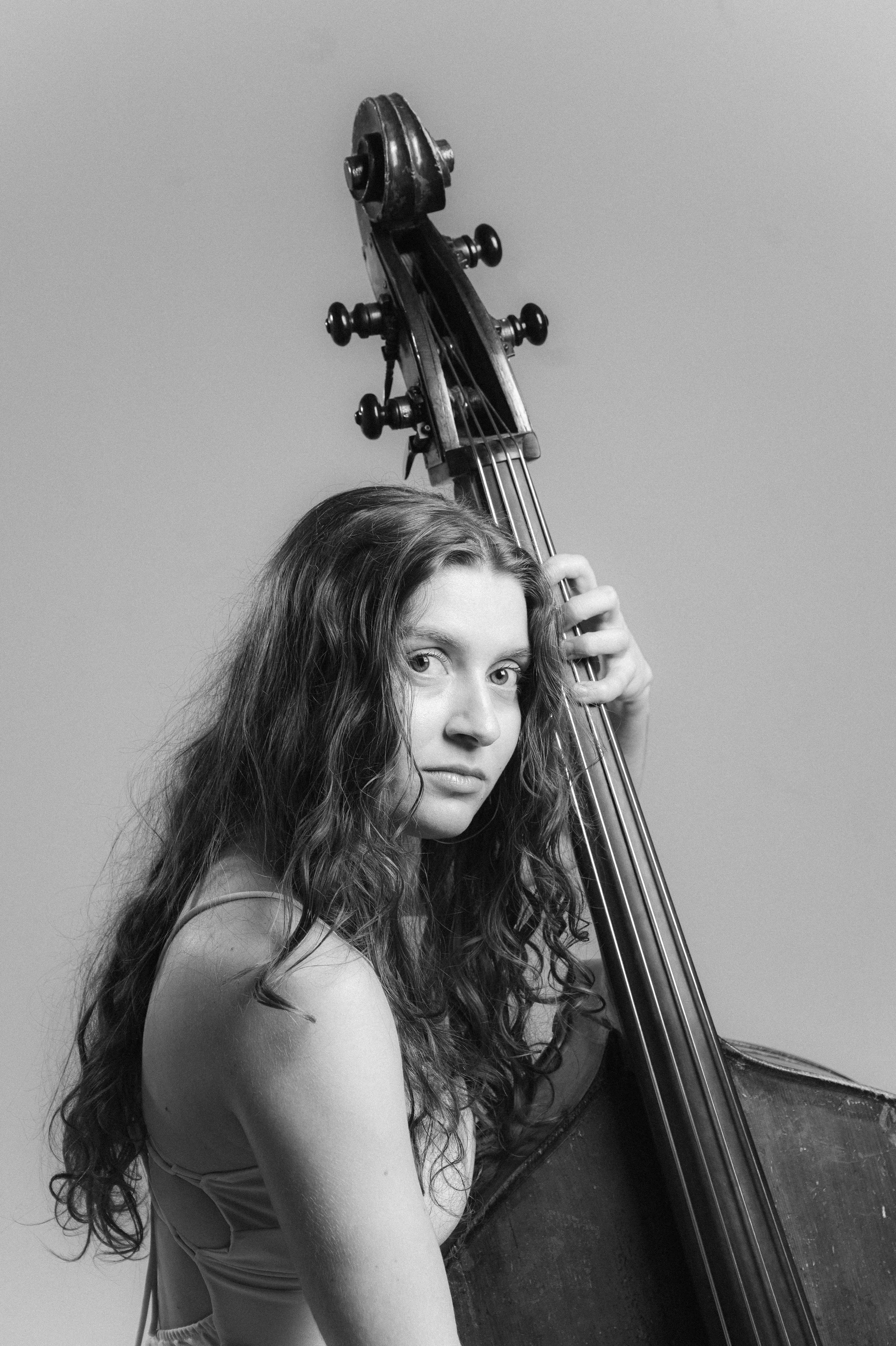 A woman with long, curly hair looks at the camera while holding a double bass upright, with her arm resting on its side, against a plain background.