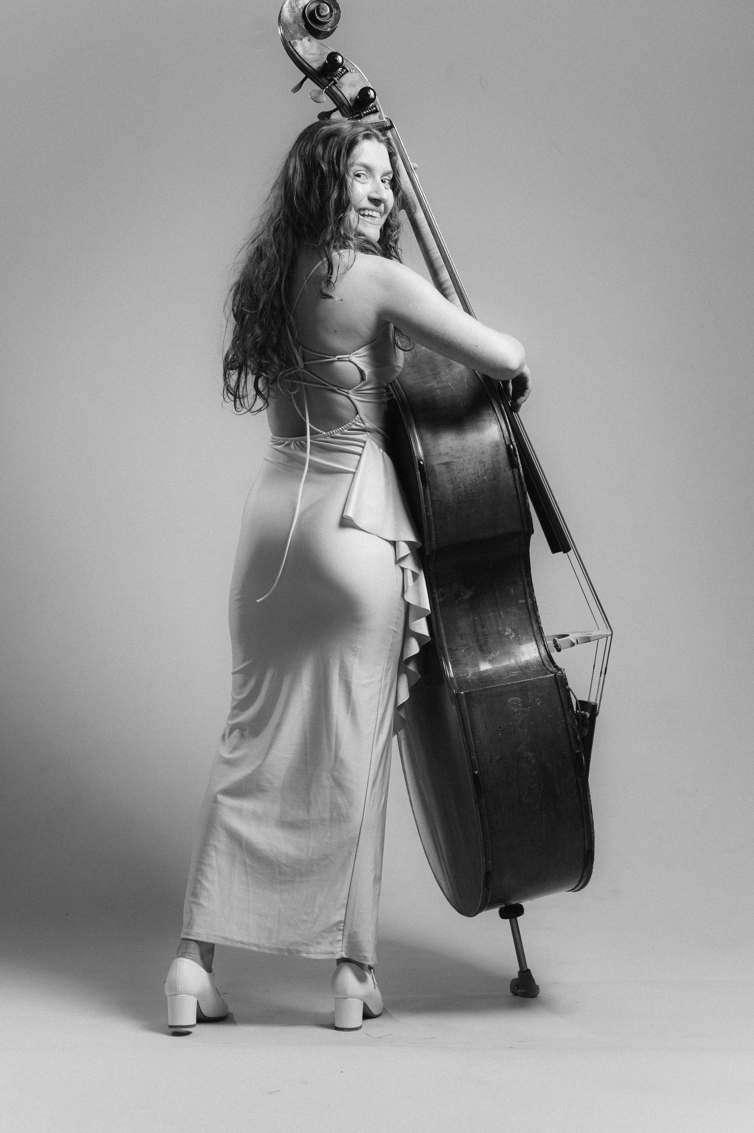 Black and white photo of a woman with long curly hair smiling and holding a double bass. She is wearing a dress with cut-out sides and high heels. The background is plain and the woman is standing, looking over her shoulder.