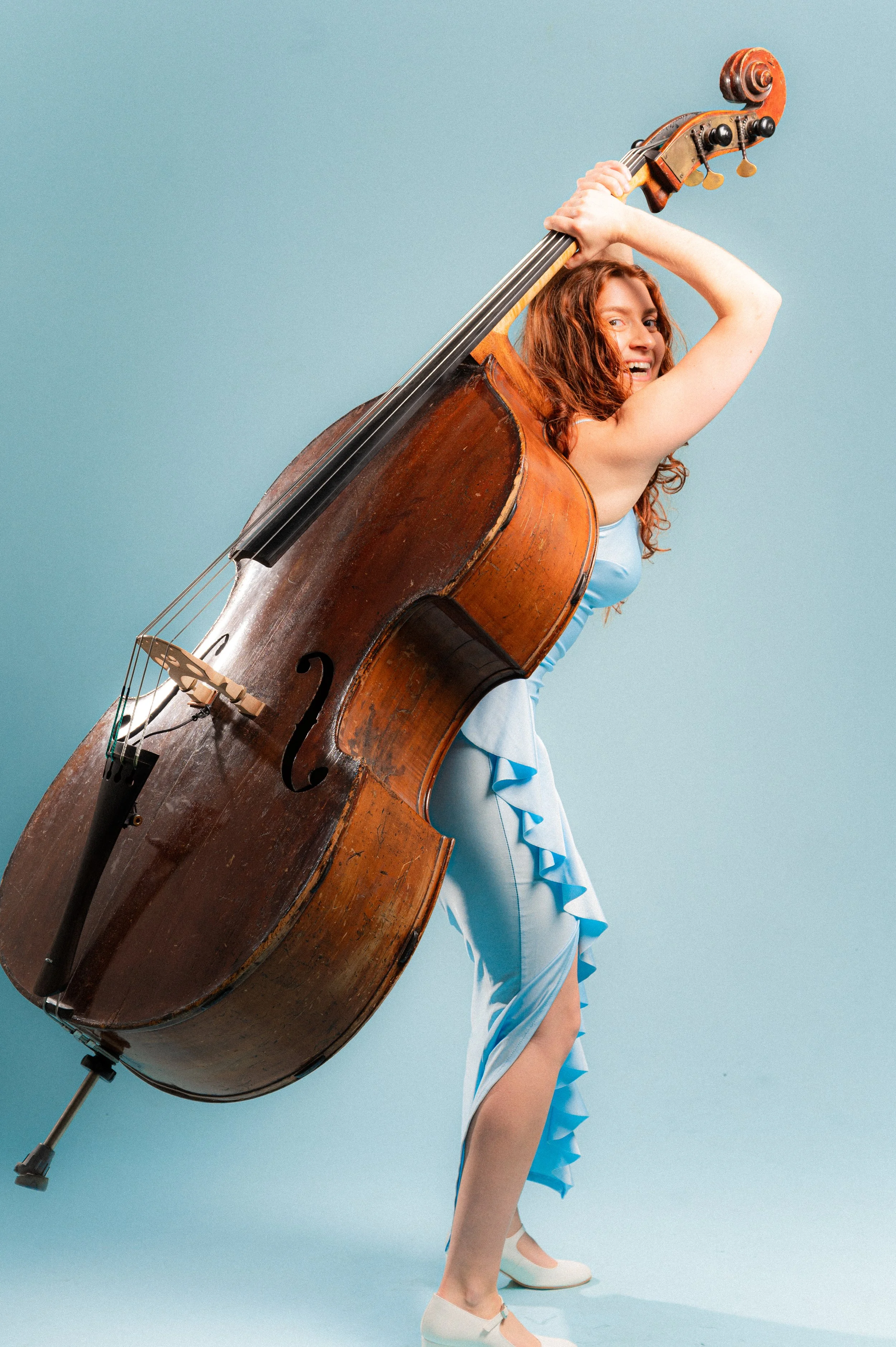 A woman with red hair, wearing a blue dress with ruffles, is holding a large, vintage wooden double bass over her shoulder and smiling. She is standing against a plain, light blue background.