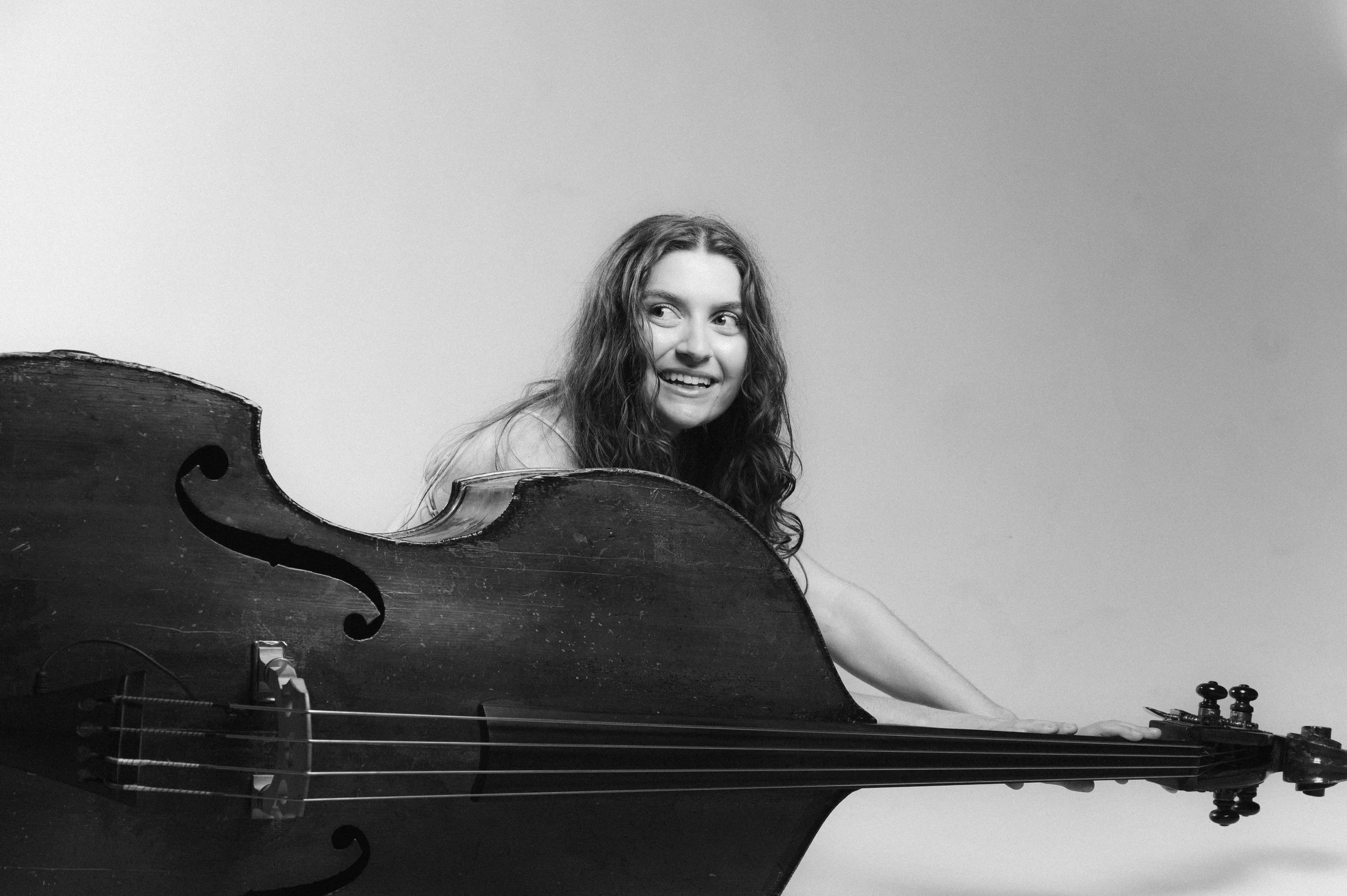 A young woman with long wavy hair smiling, partially obscured by the body of a stringed musical instrument, possibly a cello or double bass, which covers part of her face and body.