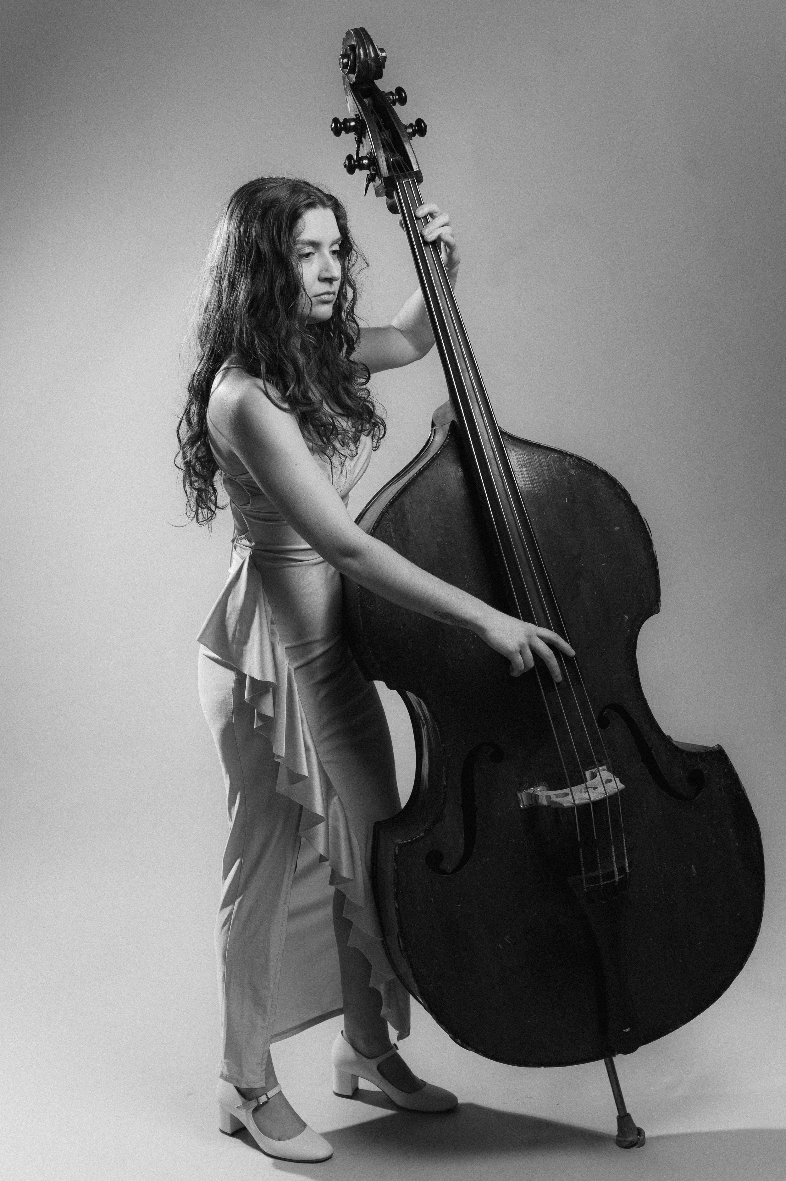 A woman with long curly hair standing and playing a double bass, dressed in elegant attire with high heels, in a studio setting with a plain background.