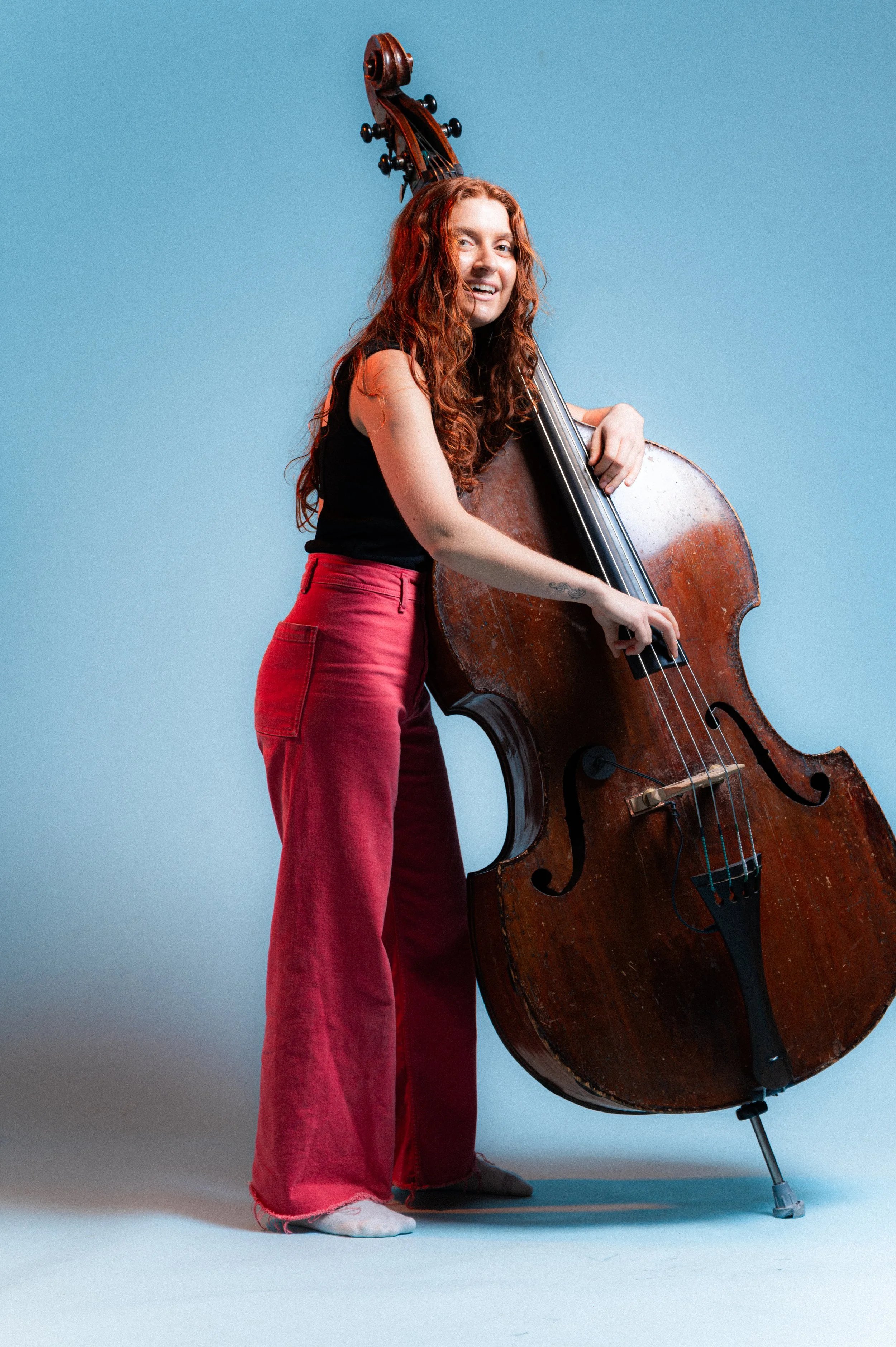 A young woman with curly red hair and a black sleeveless top playing an upright double bass against a solid blue background.