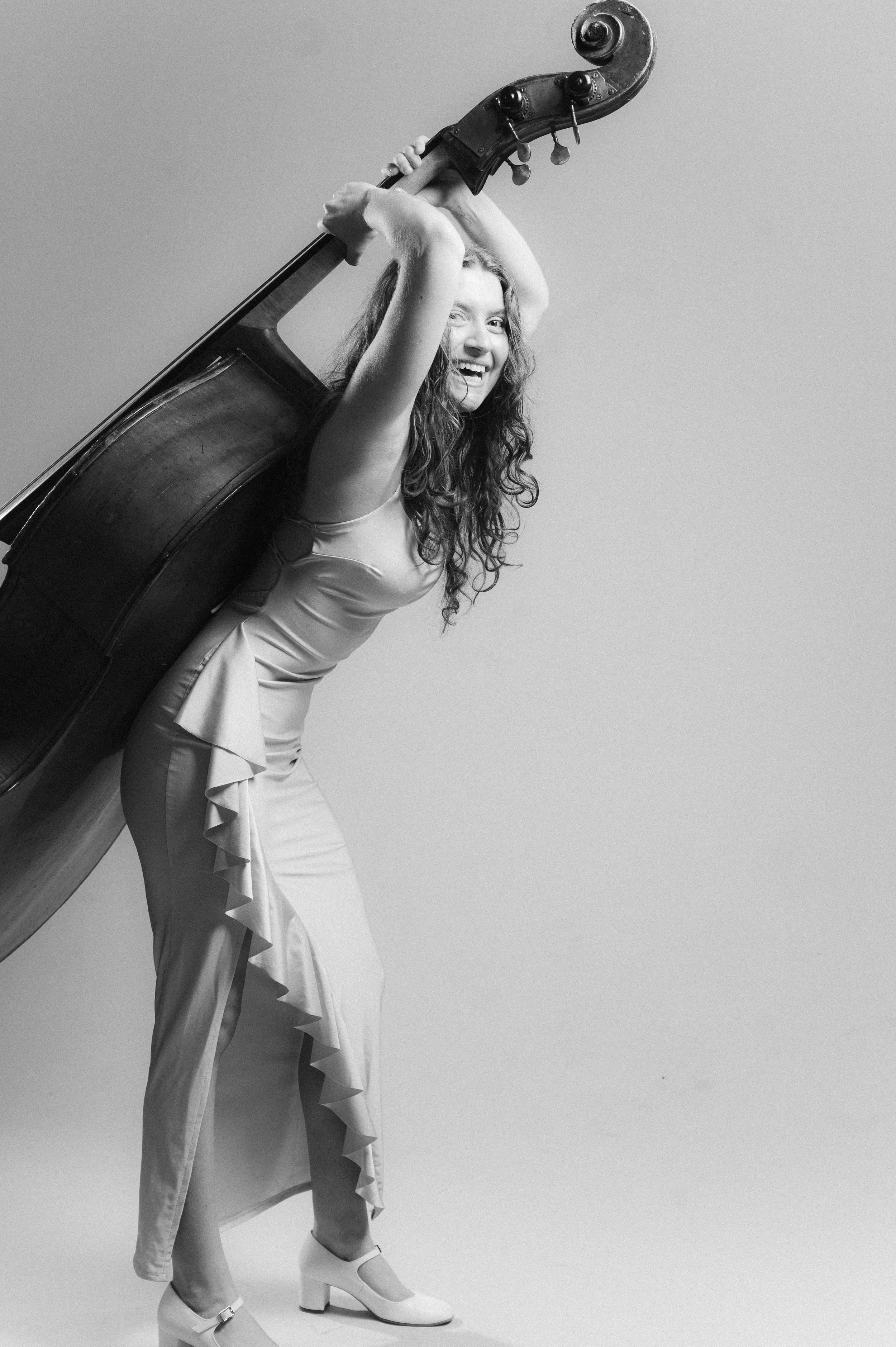 A woman in a dress, holding a double bass overhead, smiling, and standing on high-heeled shoes against a plain background.
