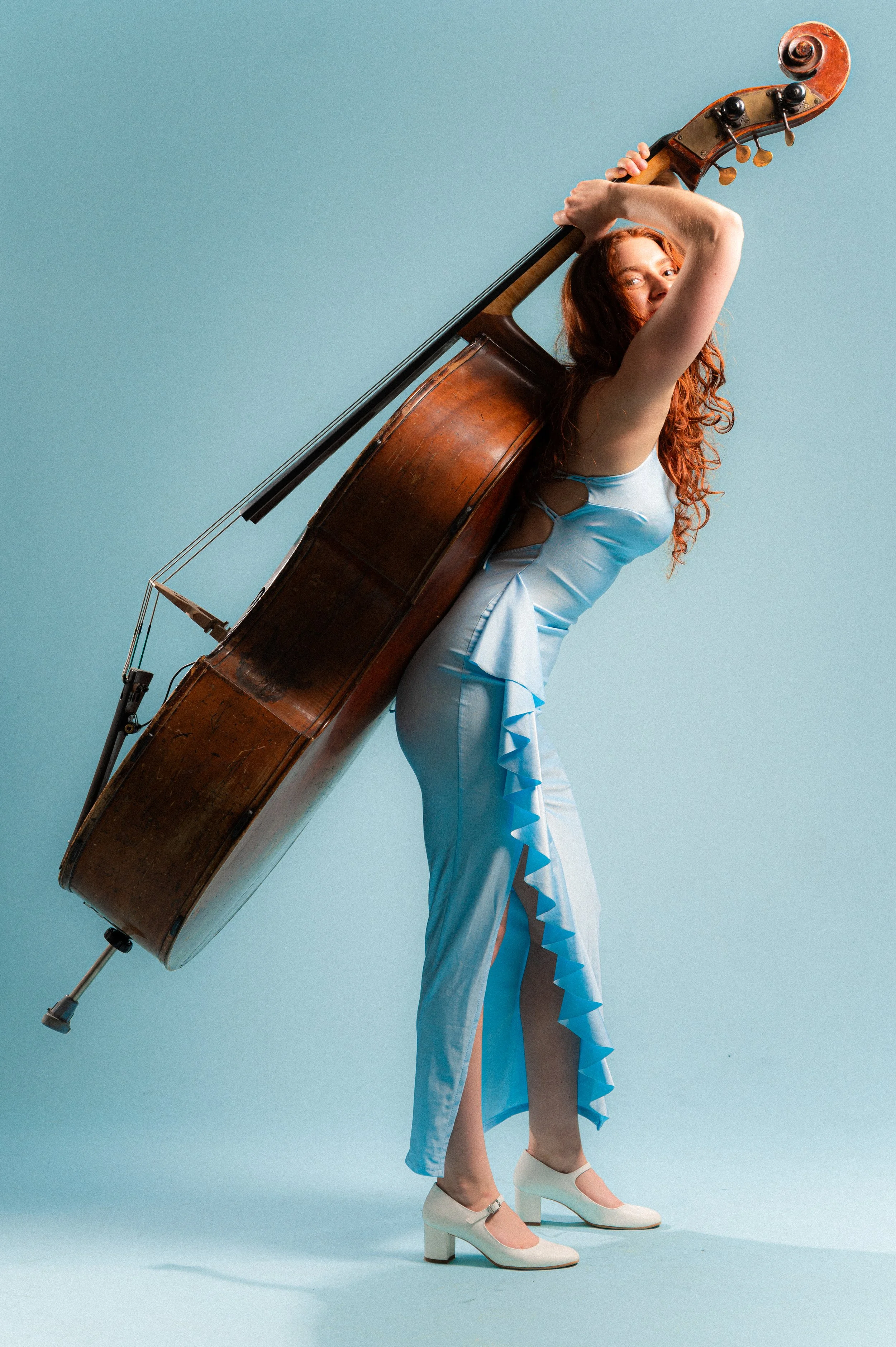 A woman with long curly red hair wearing a light blue dress and white shoes, holding a large vintage cello over her shoulder against a light blue background.