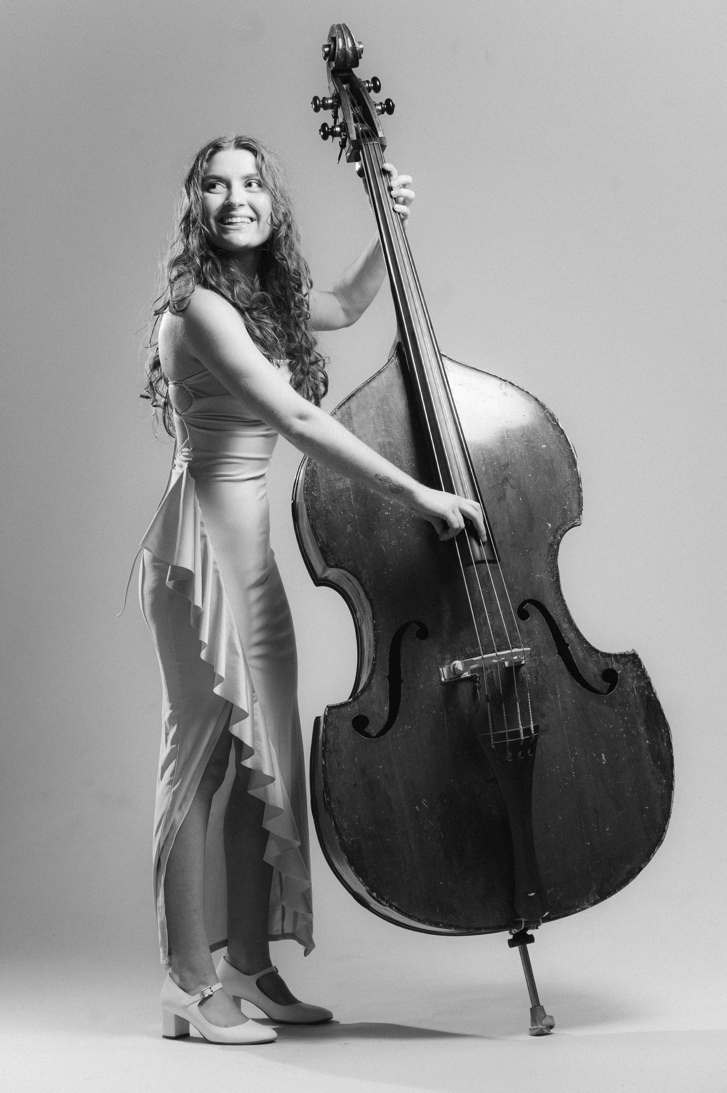 A young woman with long curly hair in a dress and high heels standing next to a large upright double bass, smiling and looking to the side, in a studio with a plain background.