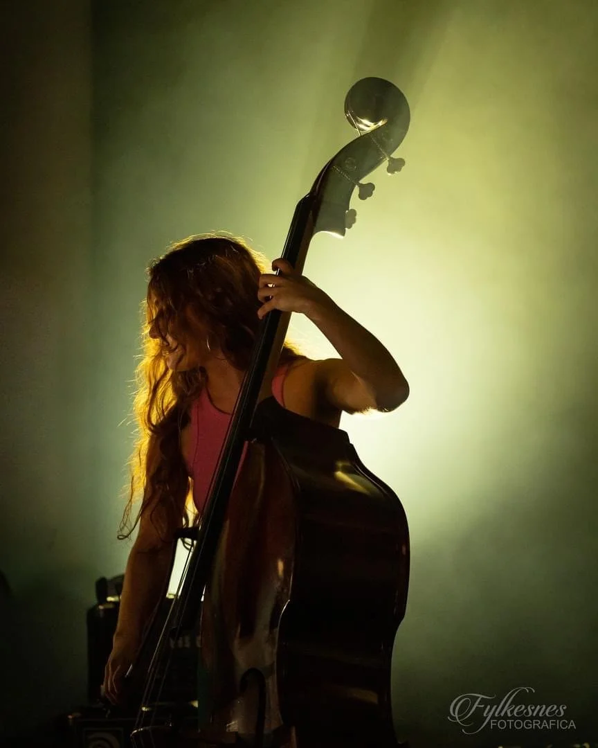 Woman with long curly hair holding a guitar in a dimly lit room with a greenish-yellow backlight.