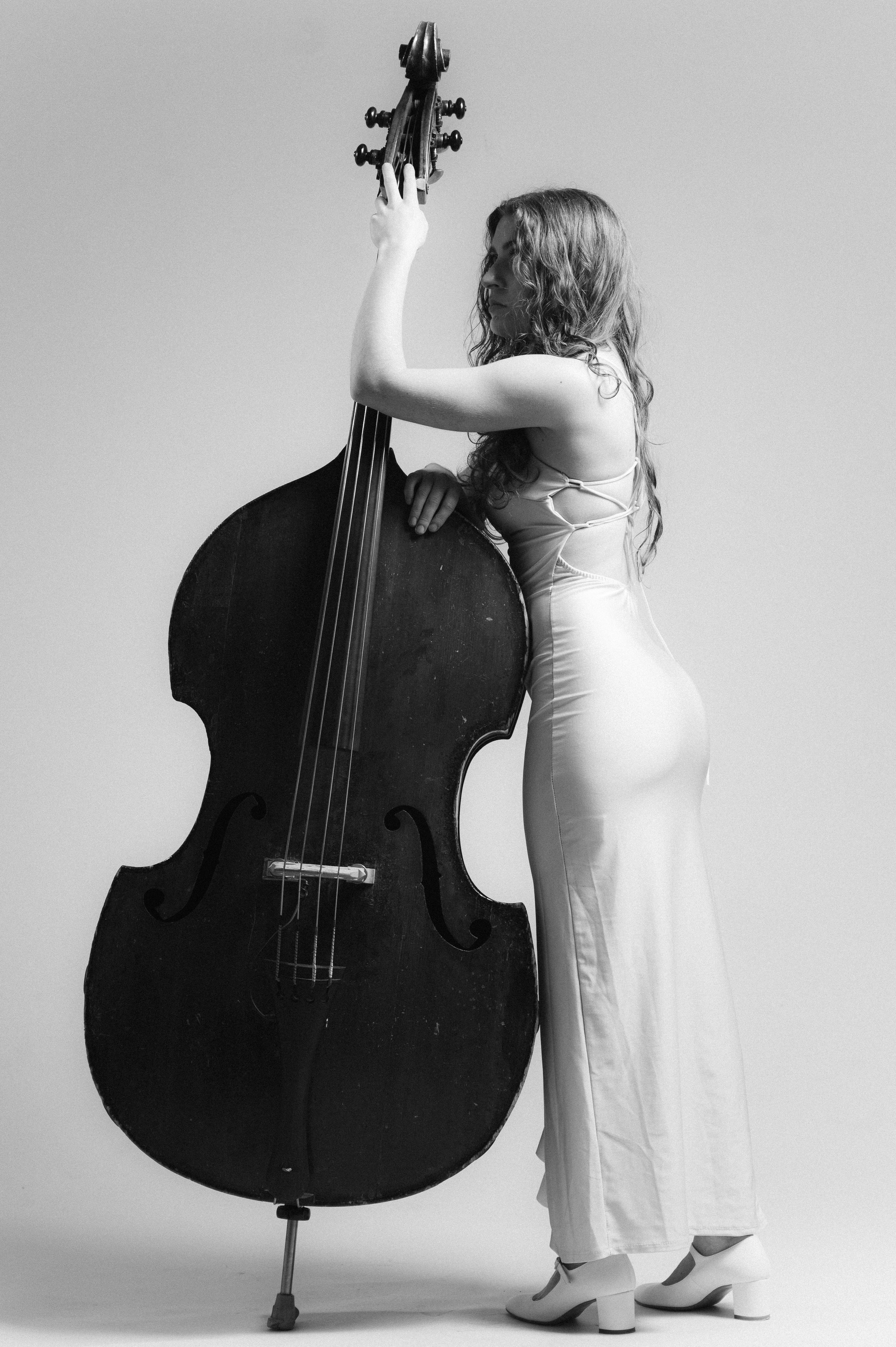 Black and white photo of a woman in a light-colored dress and high heels standing next to a large upright double bass. She is holding the tuning pegs of the instrument with one hand and looking at it, with her hair styled in loose curls. The backgrou