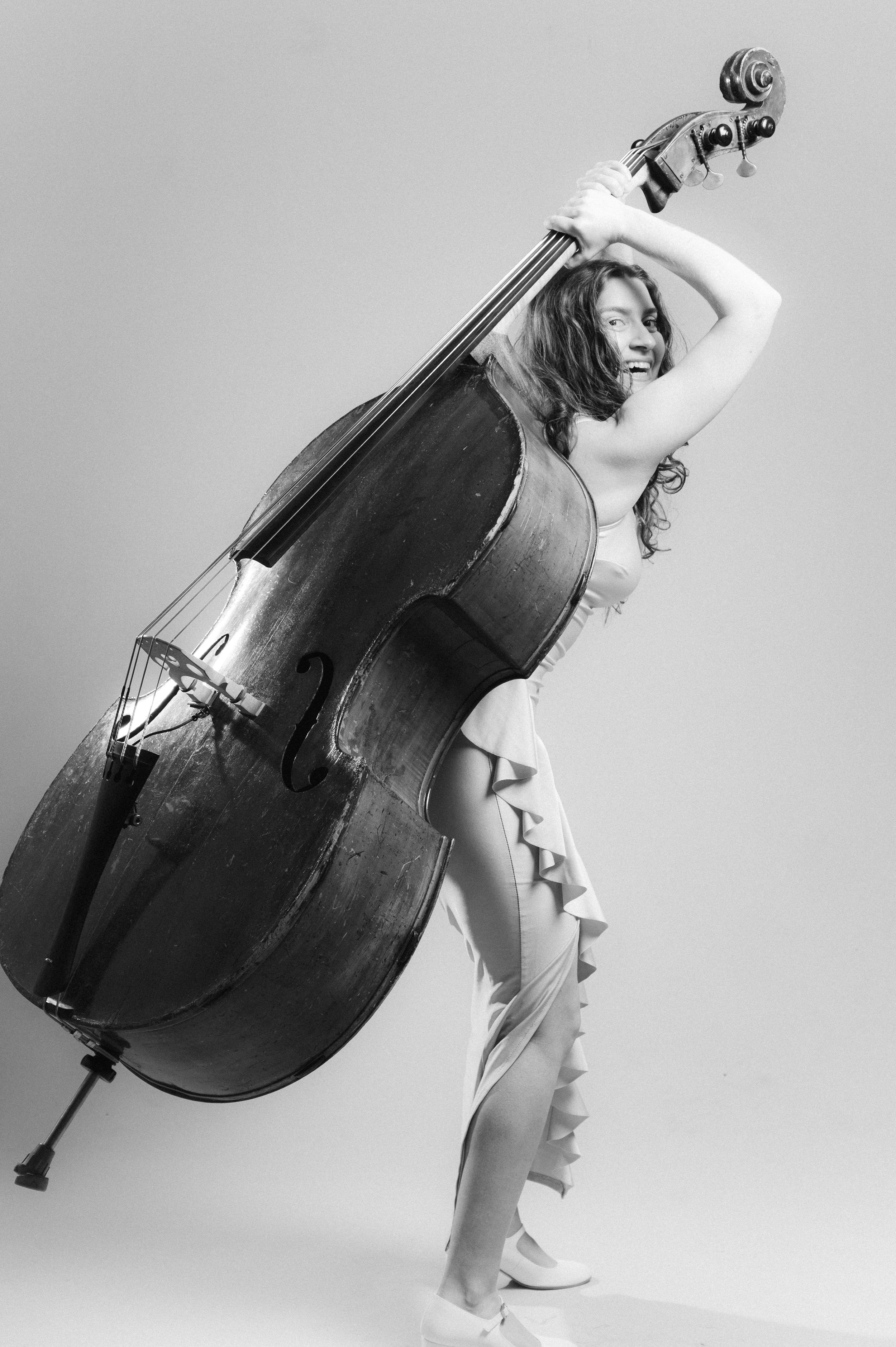 A woman in a dress and high heels holding a large, vintage double bass, smiling and leaning on it, against a plain background.