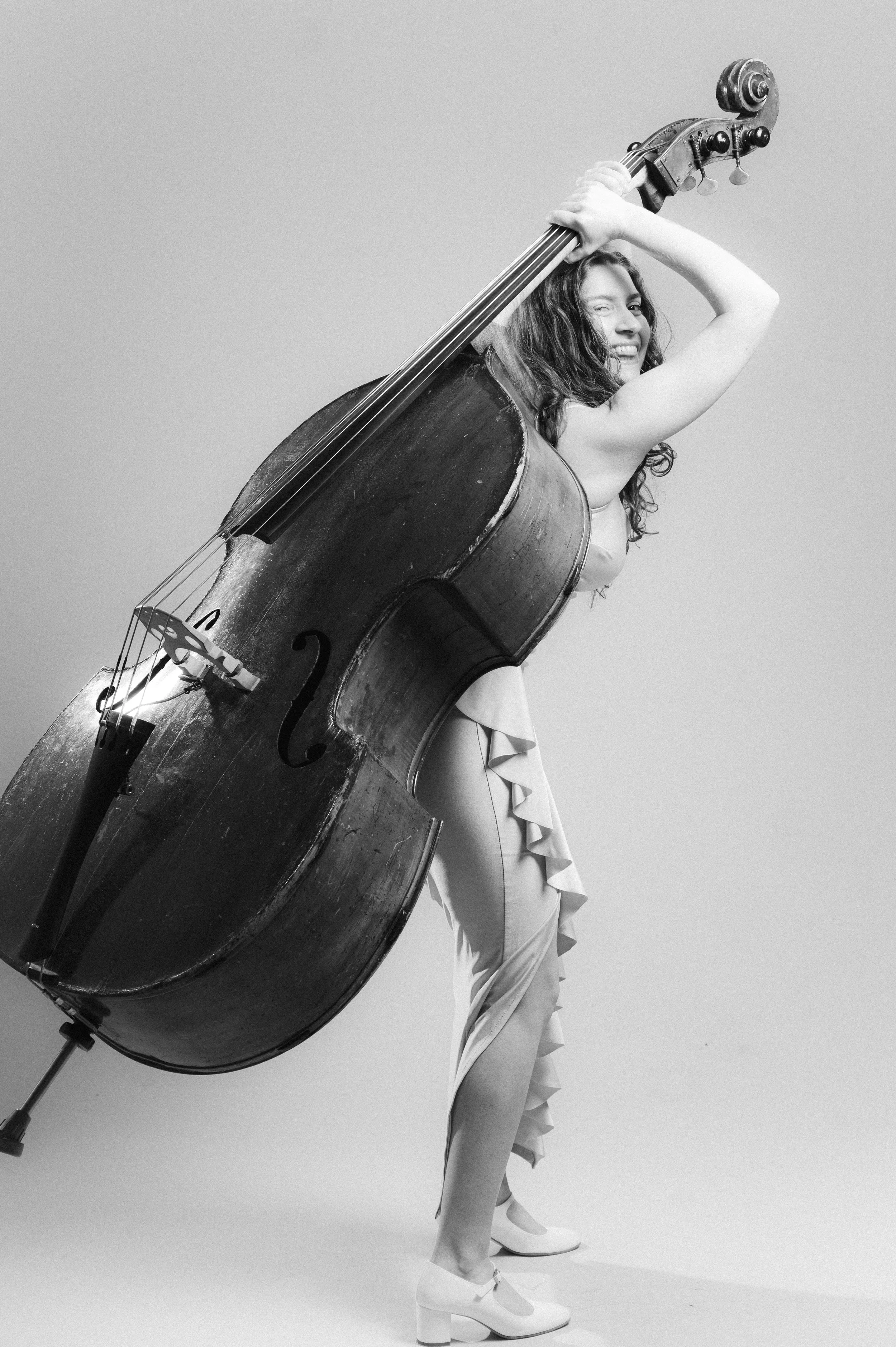 A woman with curly hair in a dress and high heels is holding a large upright double bass, smiling and looking to the side.