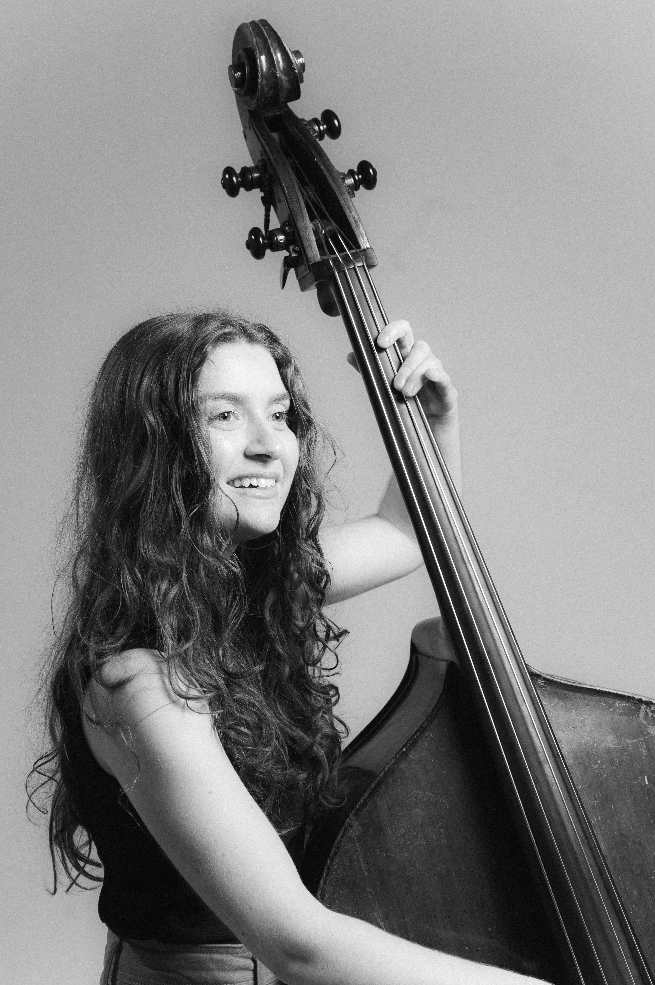 A young woman with long, wavy hair smiling and holding a double bass in a black and white photo.