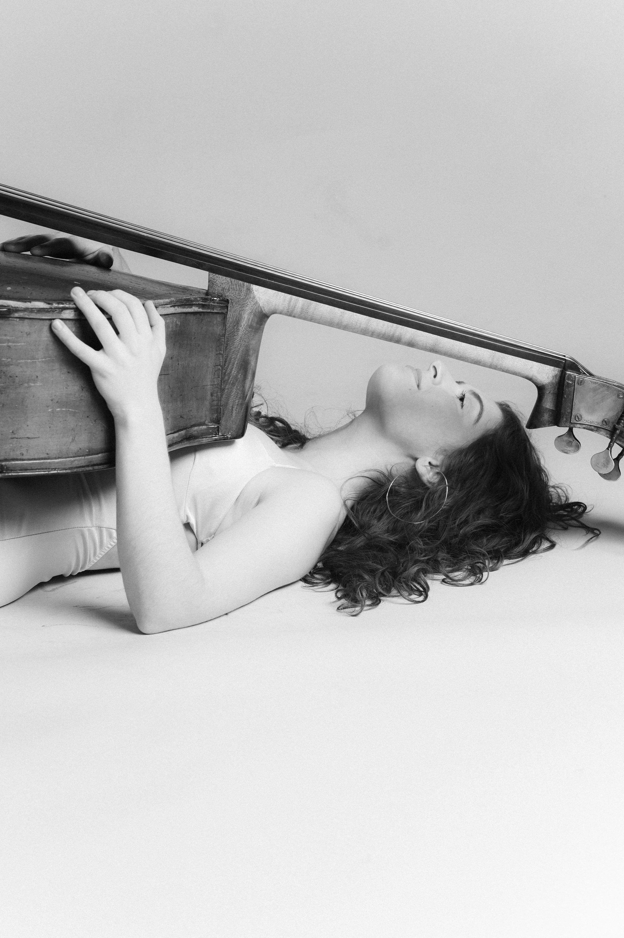 A young woman with curly hair is lying on the ground with her eyes closed, holding a violin that is positioned in front of her head on the floor.