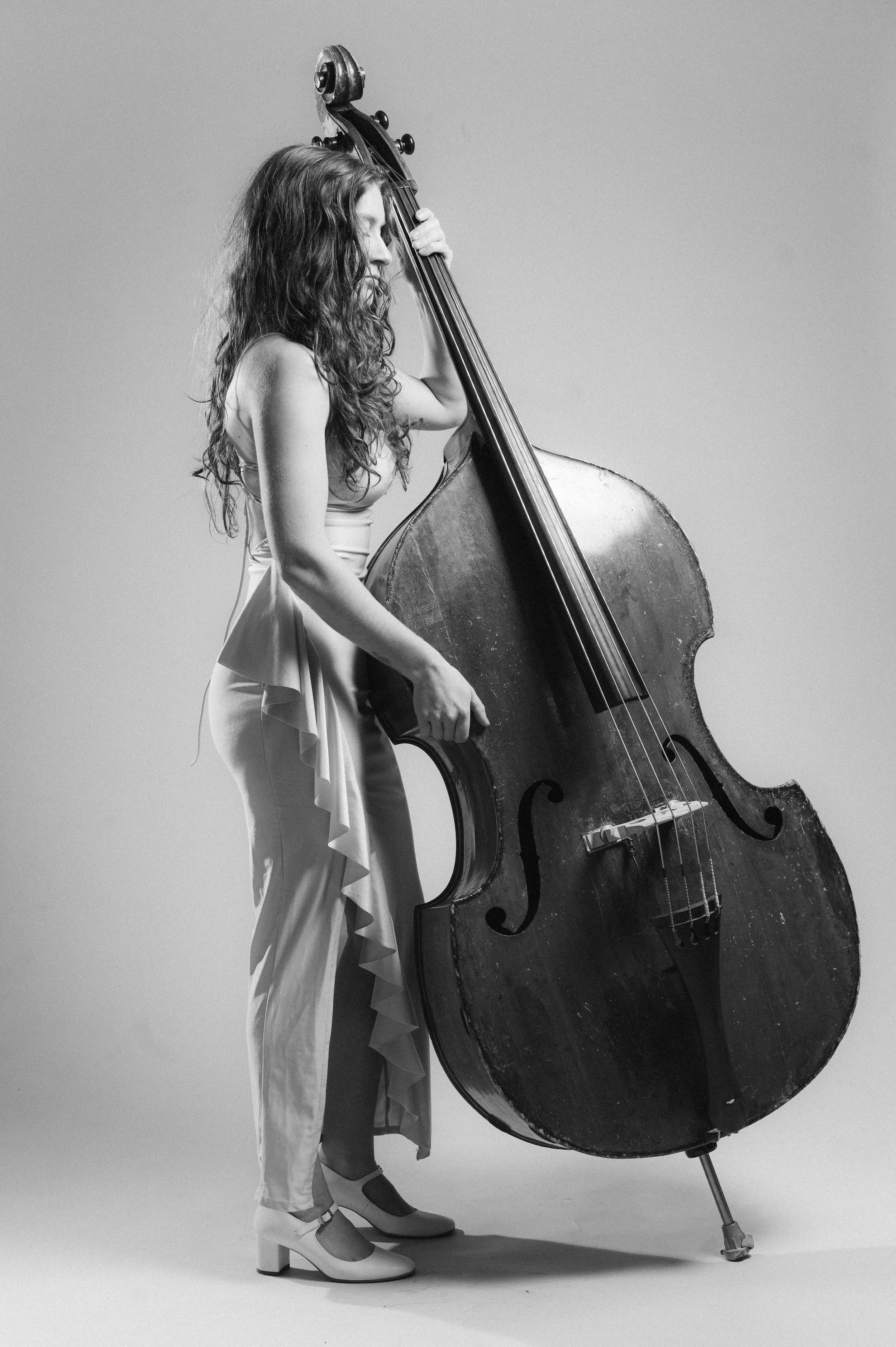A woman with wavy hair dressed in a light-colored ruffled dress and platform shoes stands with her eyes closed, gently resting her head on a large upright double bass. The instrument has a worn wooden body and is supported by a metal stand, contrasti