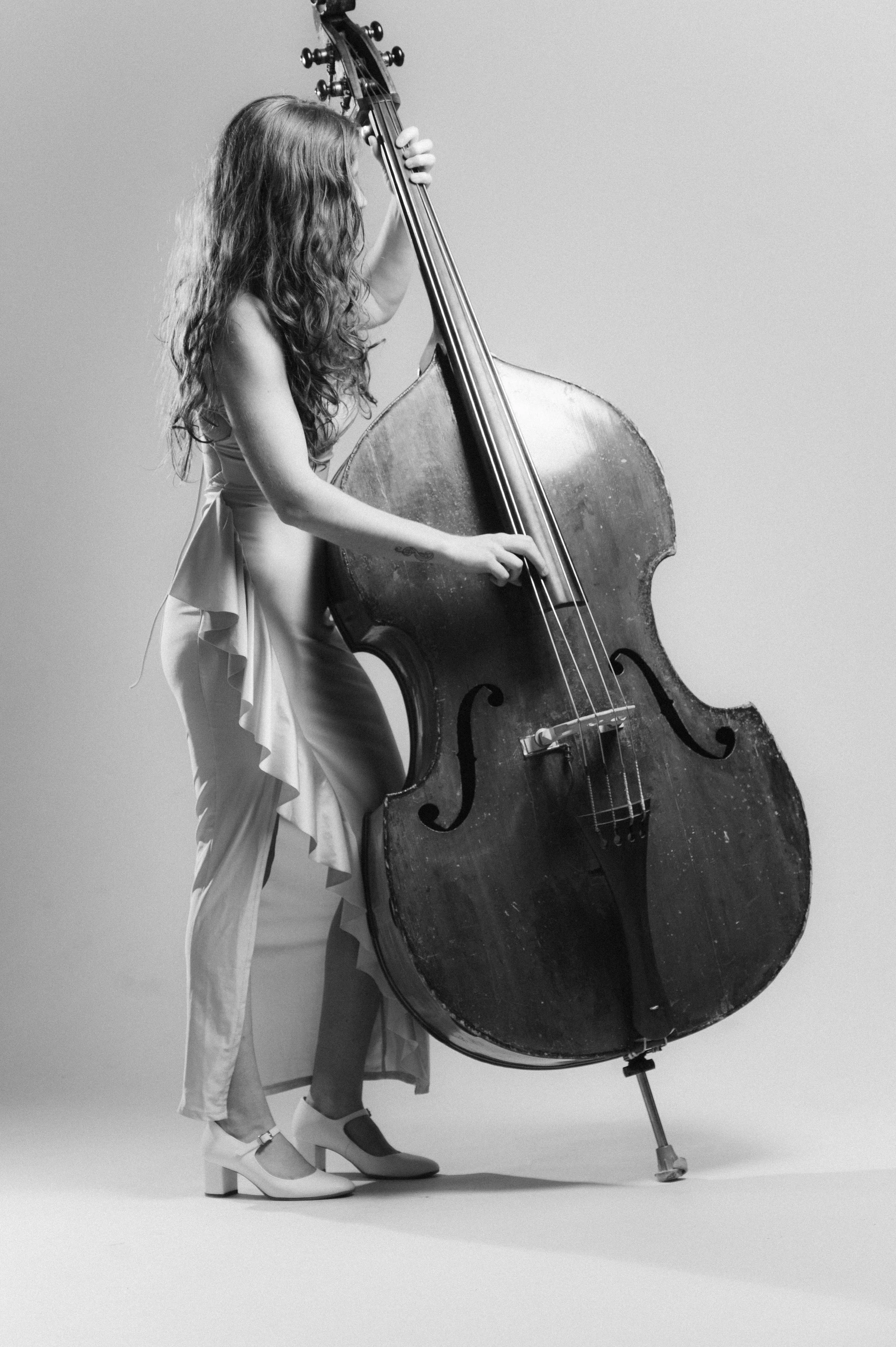 Black and white photo of a woman with long wavy hair playing a double bass. She wears a dress with ruffled layers and shoes with a chunky heel. The background is plain.