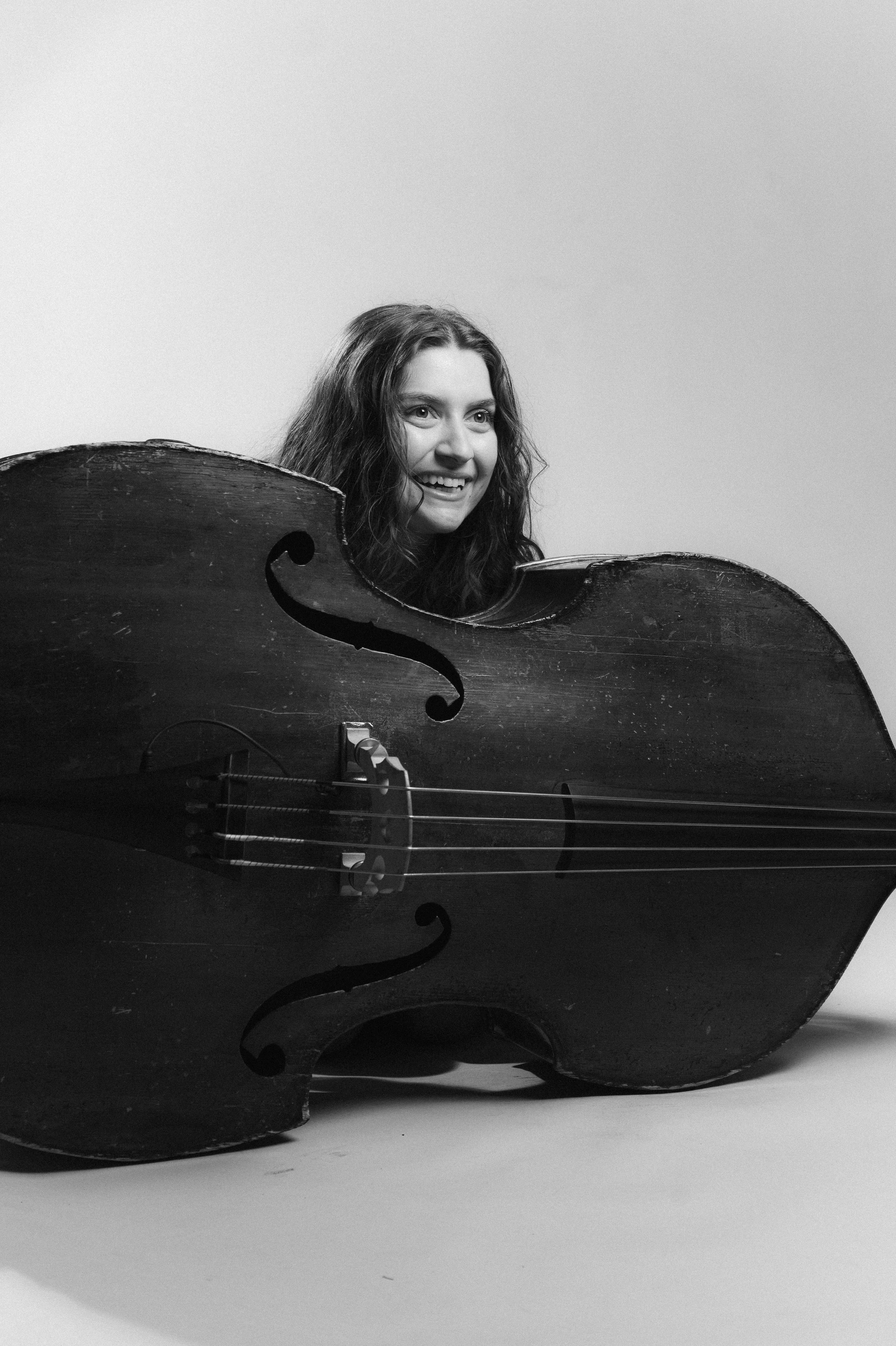Black and white photo of a young woman with curly hair, smiling, partially hidden behind a large musical instrument, likely a double bass, with a plain light-colored background.