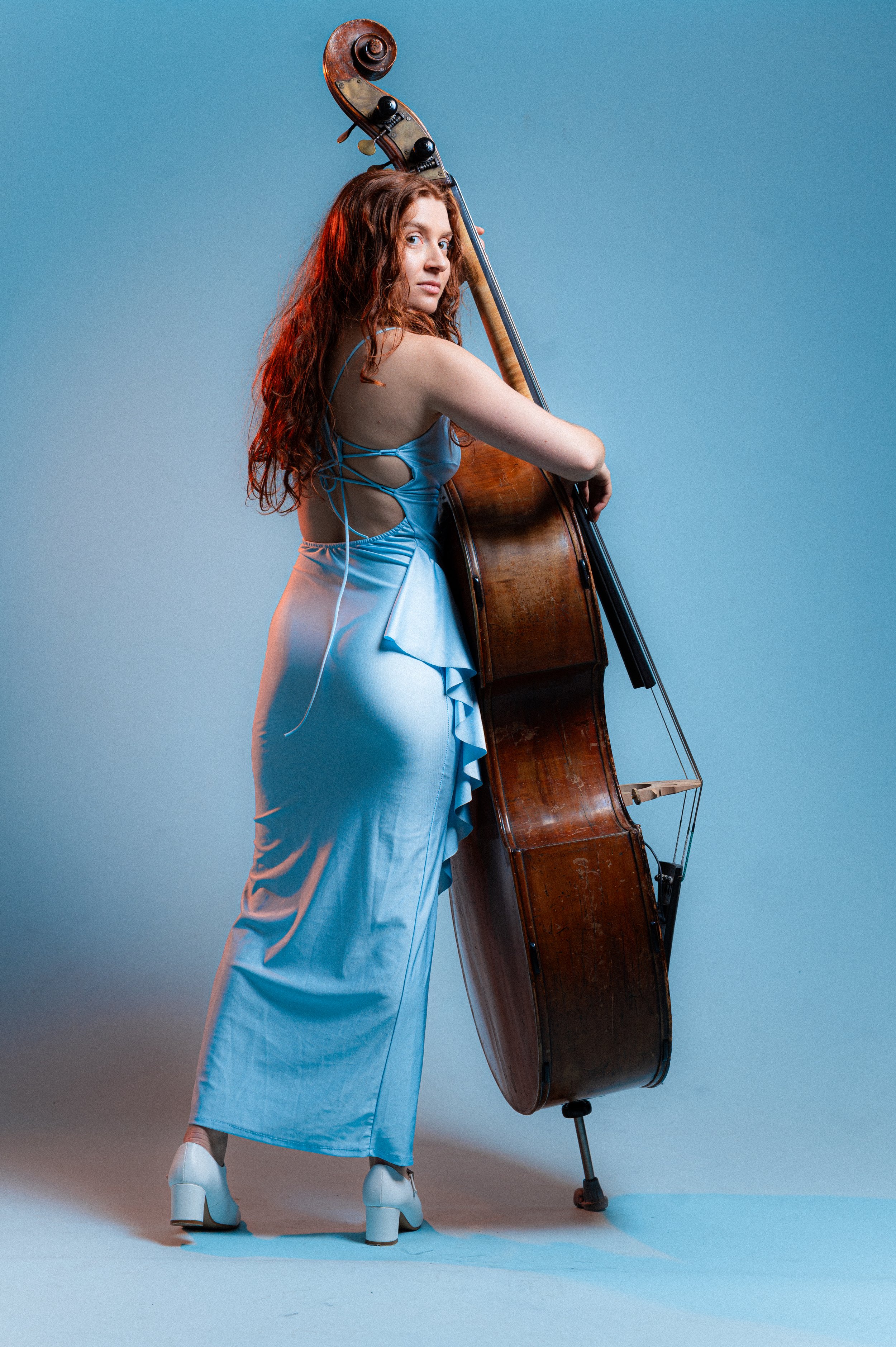 A woman with long curly red hair wearing a light blue satin dress and high heels, standing against a plain background, holding a large upright bass. The bass has a wooden body and strings, with some hand and finger positioning indicating she is playi