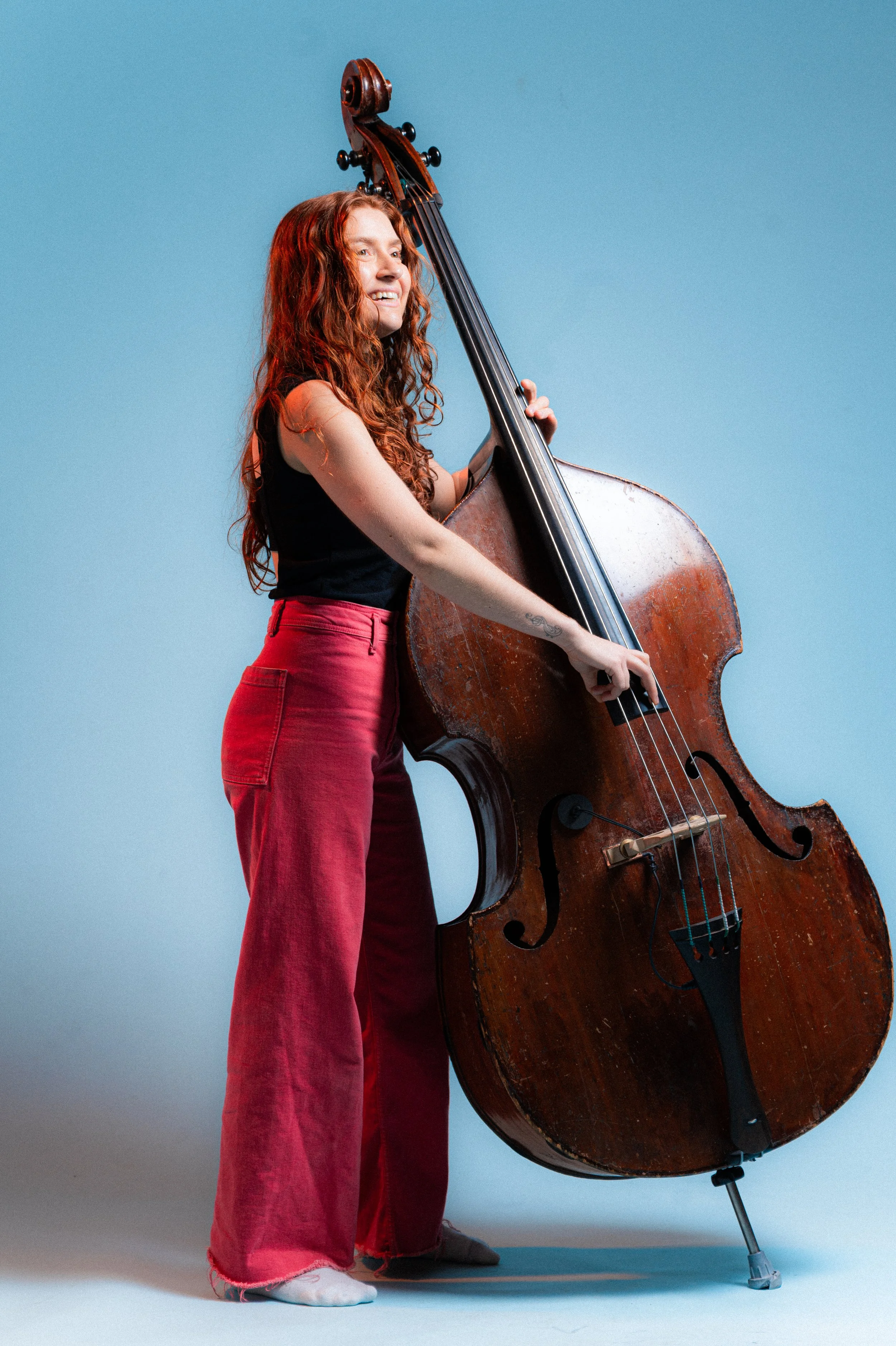 A young woman with long, curly red hair wearing red pants and a black tank top, standing on a light blue background, holding a large vintage double bass upright and smiling.