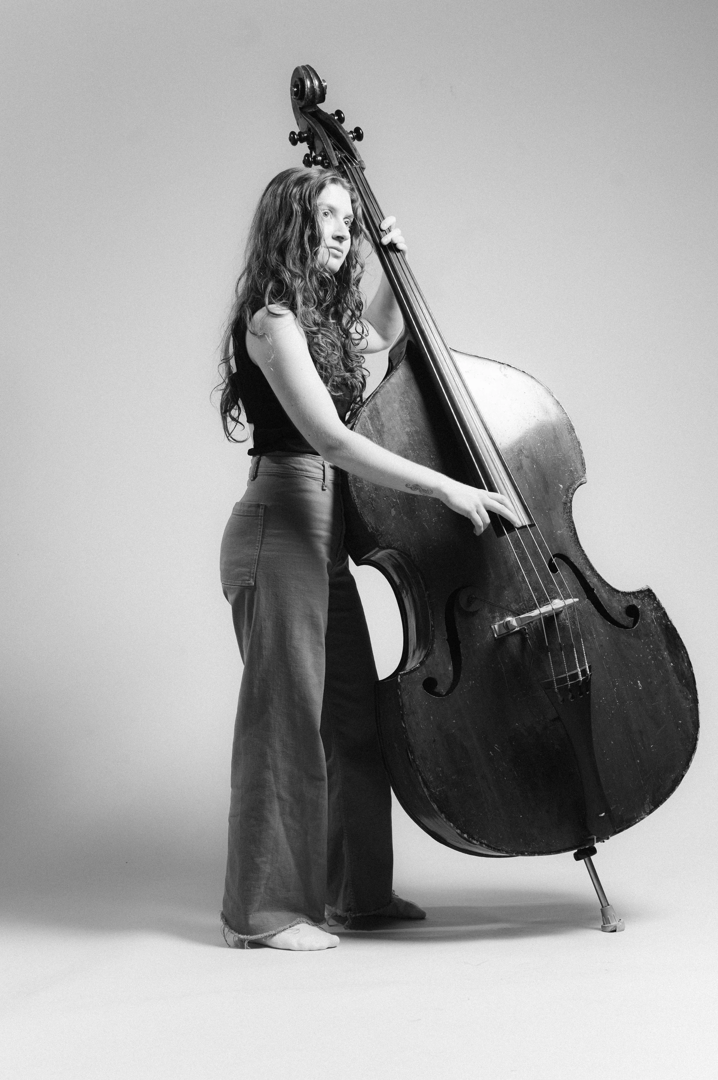 A woman with long curly hair standing and playing a double bass against a plain background.