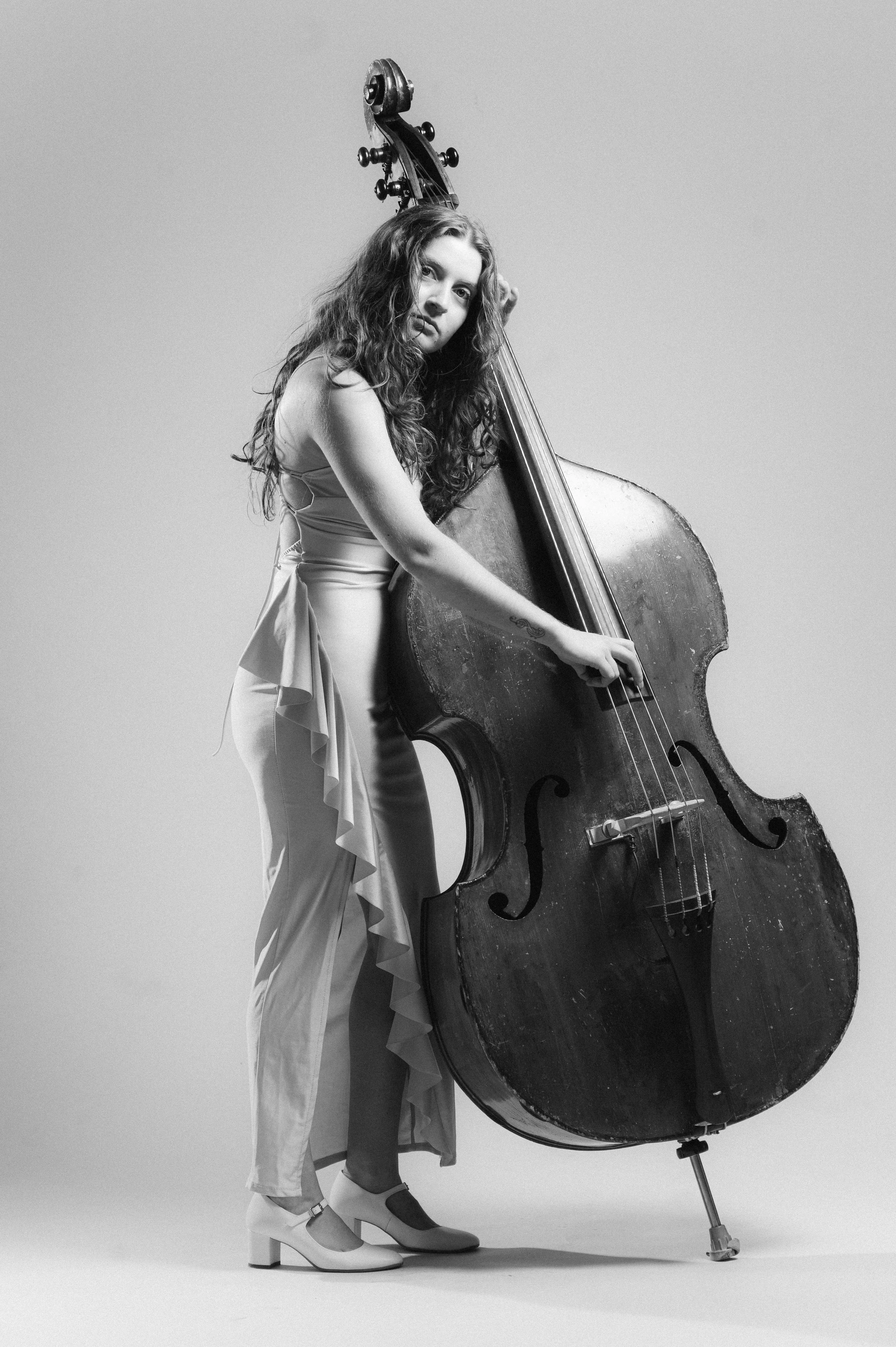 A woman with long curly hair in a light dress and high heels is standing next to a large upright bass, holding it with one hand. The image is in black and white against a plain background.