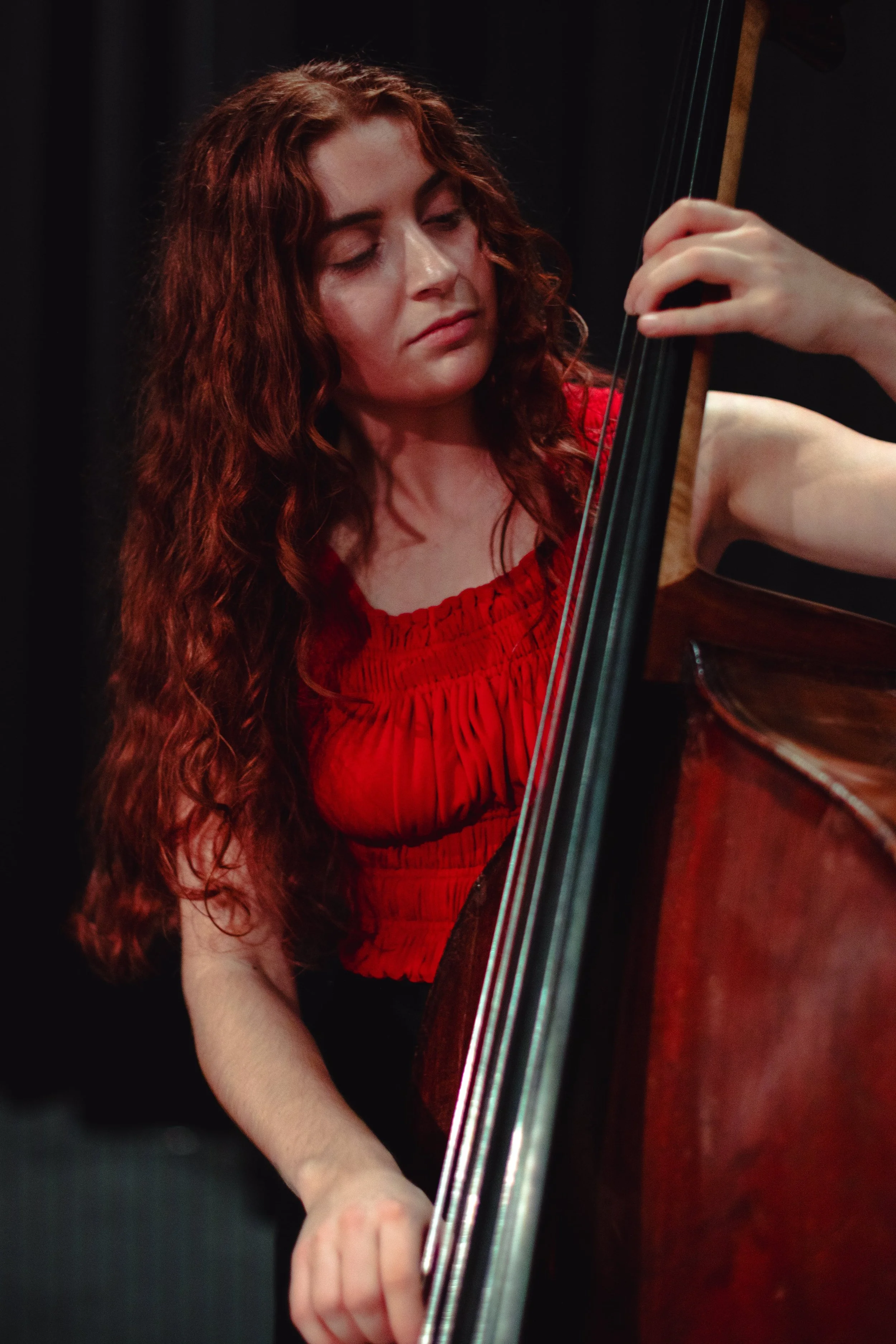 A woman with long, curly red hair playing a upright bass string instrument, wearing a red dress with pleated details.