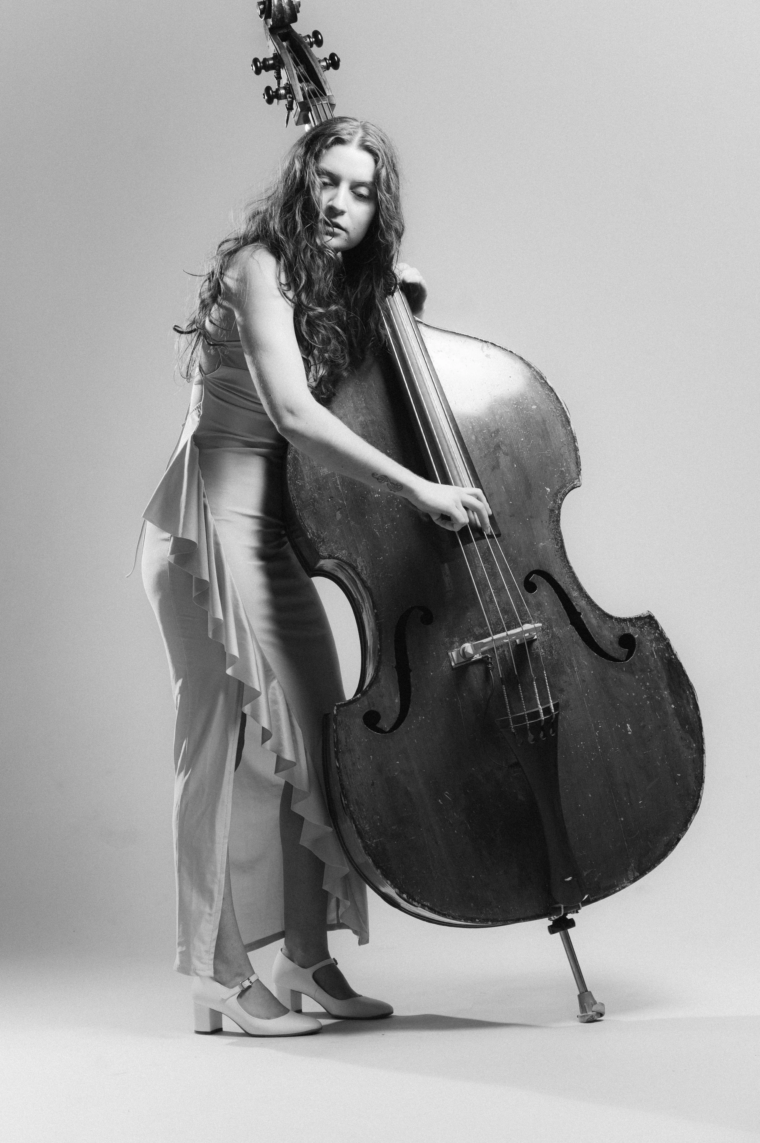 Black and white photo of a woman with curly hair playing a double bass, standing in a studio with plain background, wearing a dress and high heels.