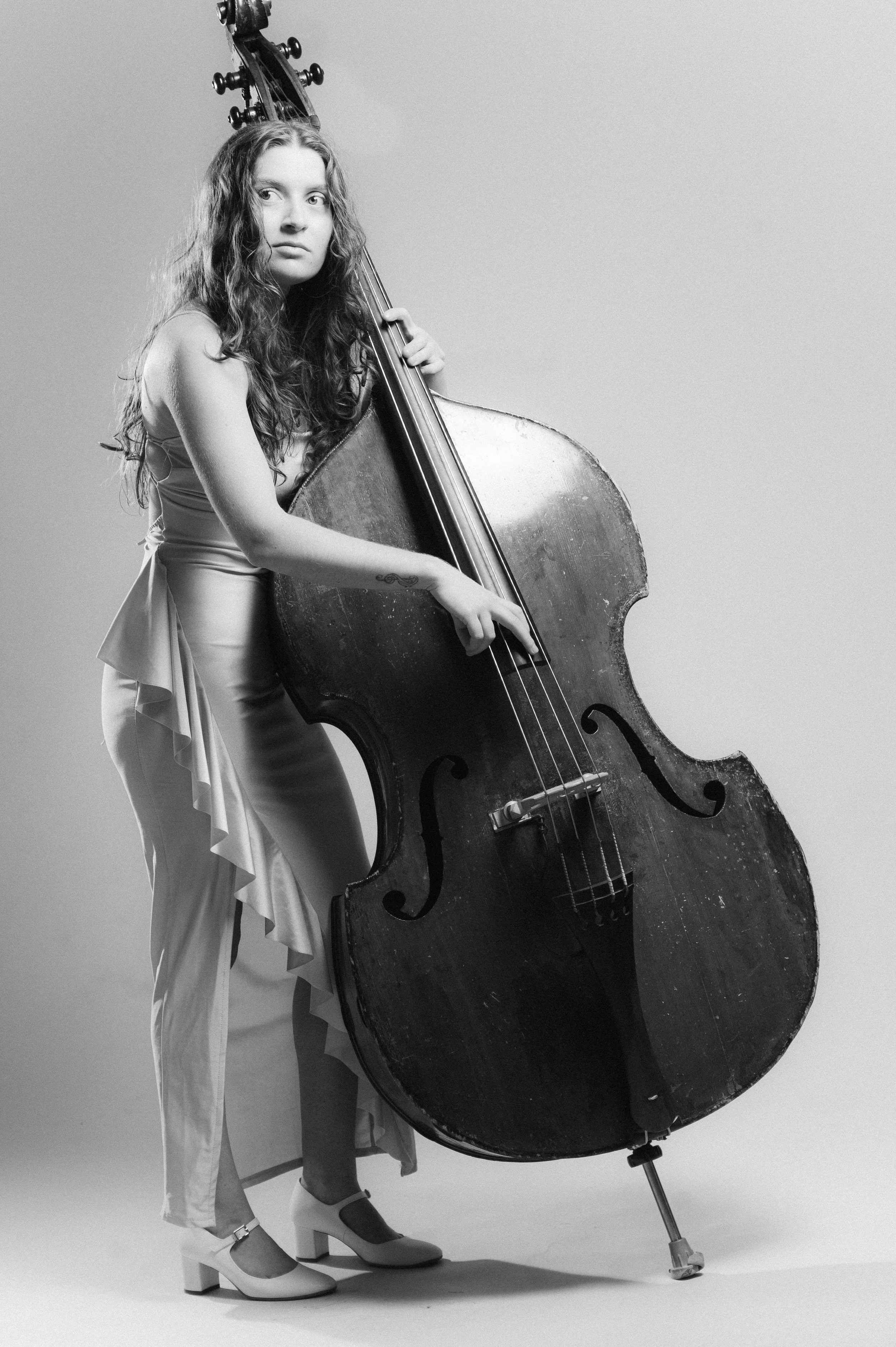 A woman with long curly hair wearing a sleeveless top, light-colored pants with ruffles on the sides, and high-heeled shoes, stands next to a large upright double bass. She is holding the instrument with one hand and looking slightly to the side.
