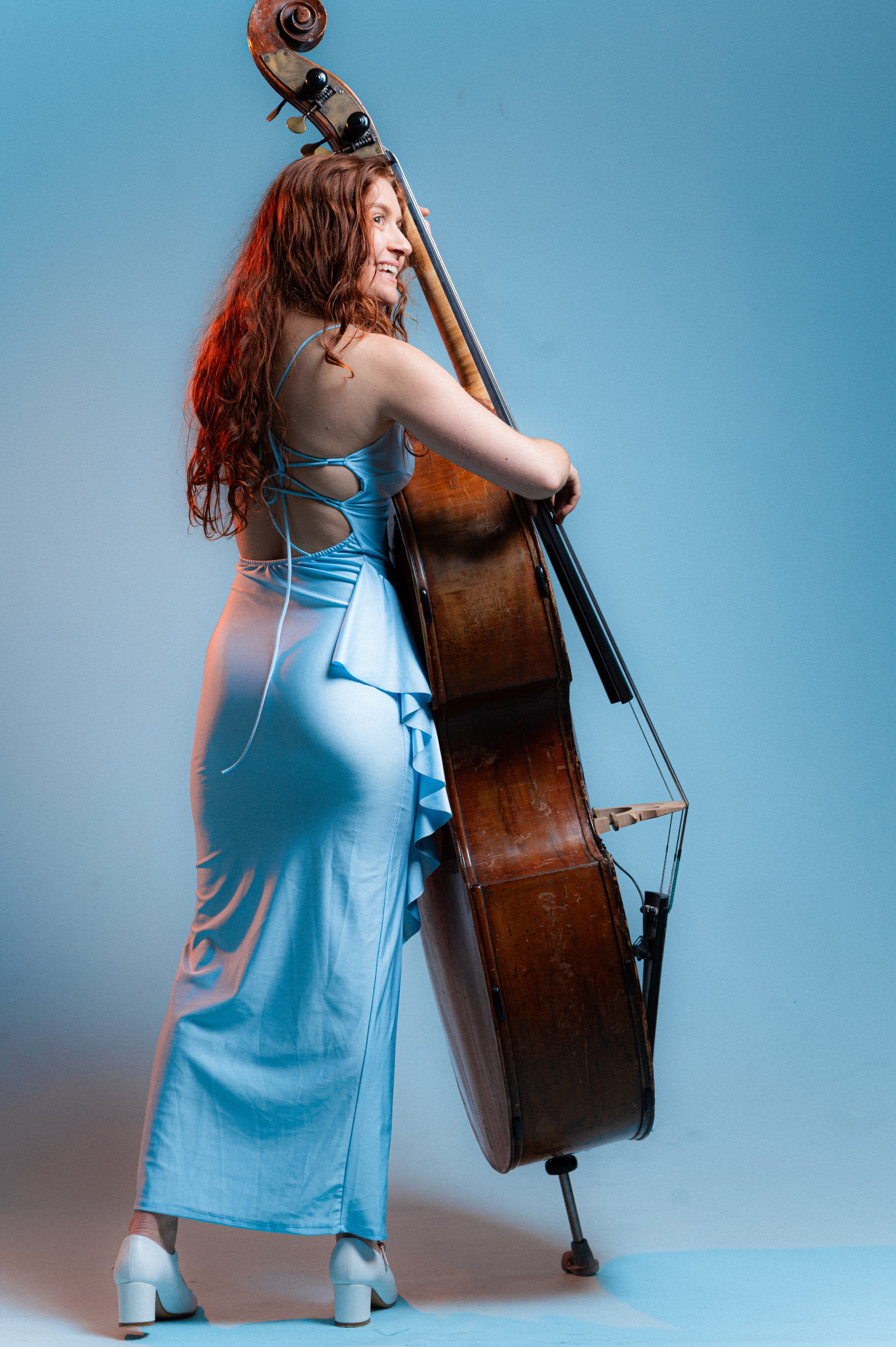 A woman with long, wavy red hair in a shiny light blue dress with open back and high heels stands next to a double bass against a plain blue background, smiling and looking to her left.