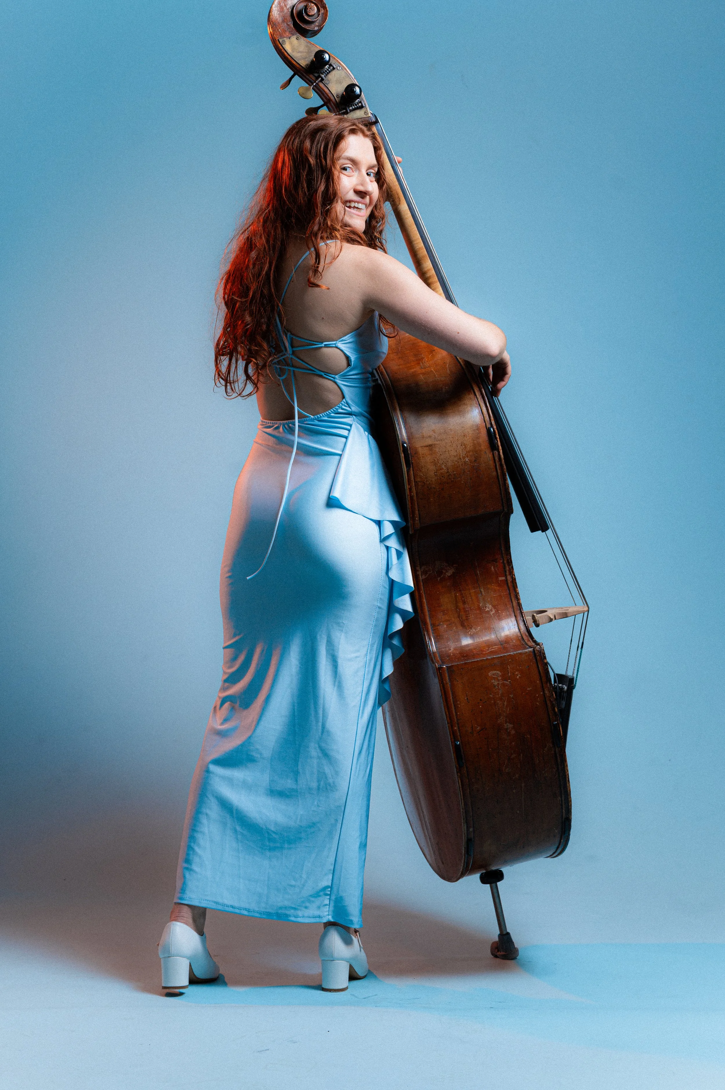 A woman with long curly red hair, wearing a light blue satin dress, white high heels, and recording earphones, is posing with a large upright wooden double bass.