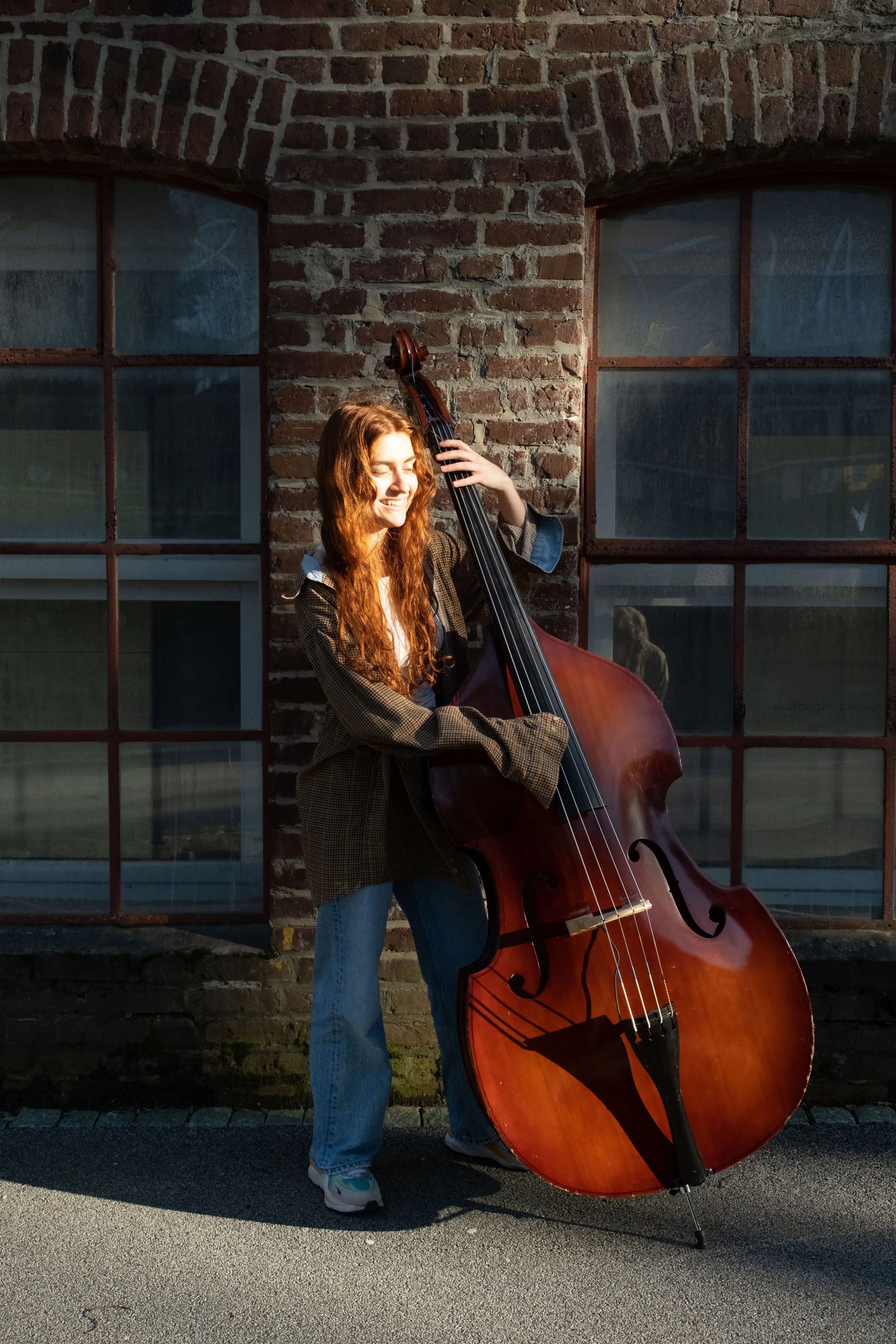 A young woman with long red hair is playing a double bass outdoors in front of a brick building with large windows, smiling with her eyes closed.