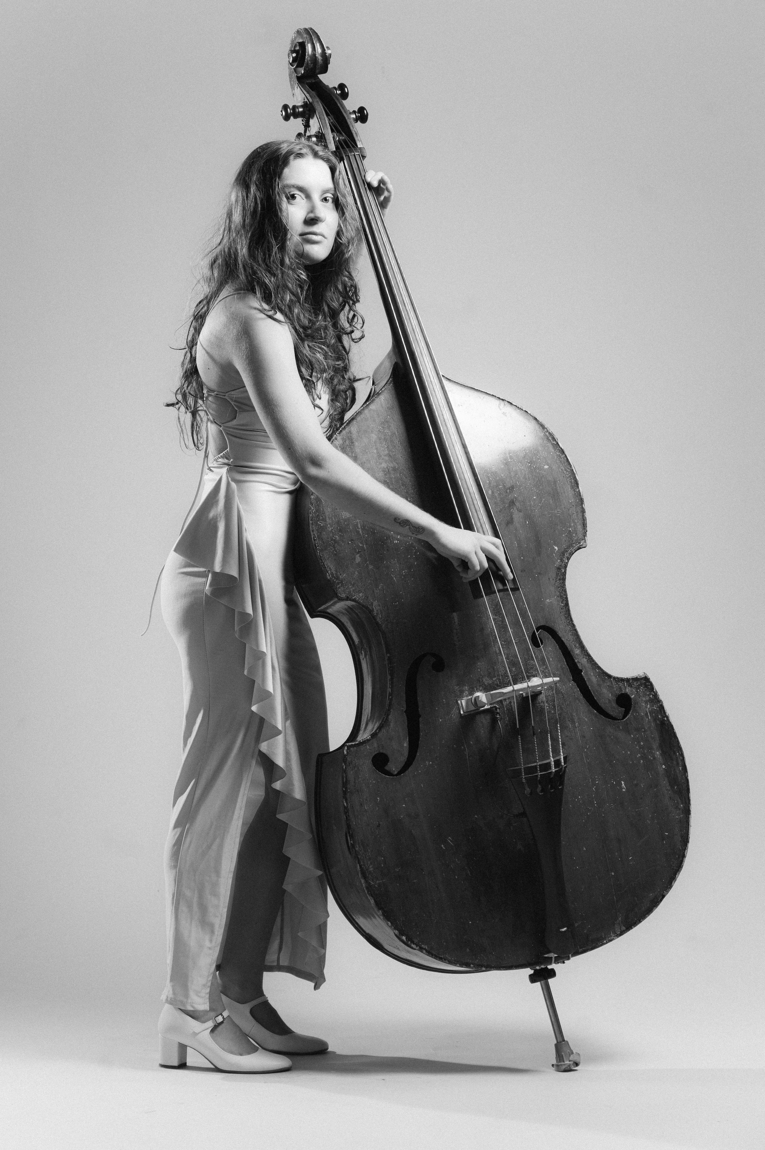 A young woman with long, curly hair in a formal dress, wearing heels, standing beside a large upright double bass, holding its neck, in a professional studio setting with a plain background.