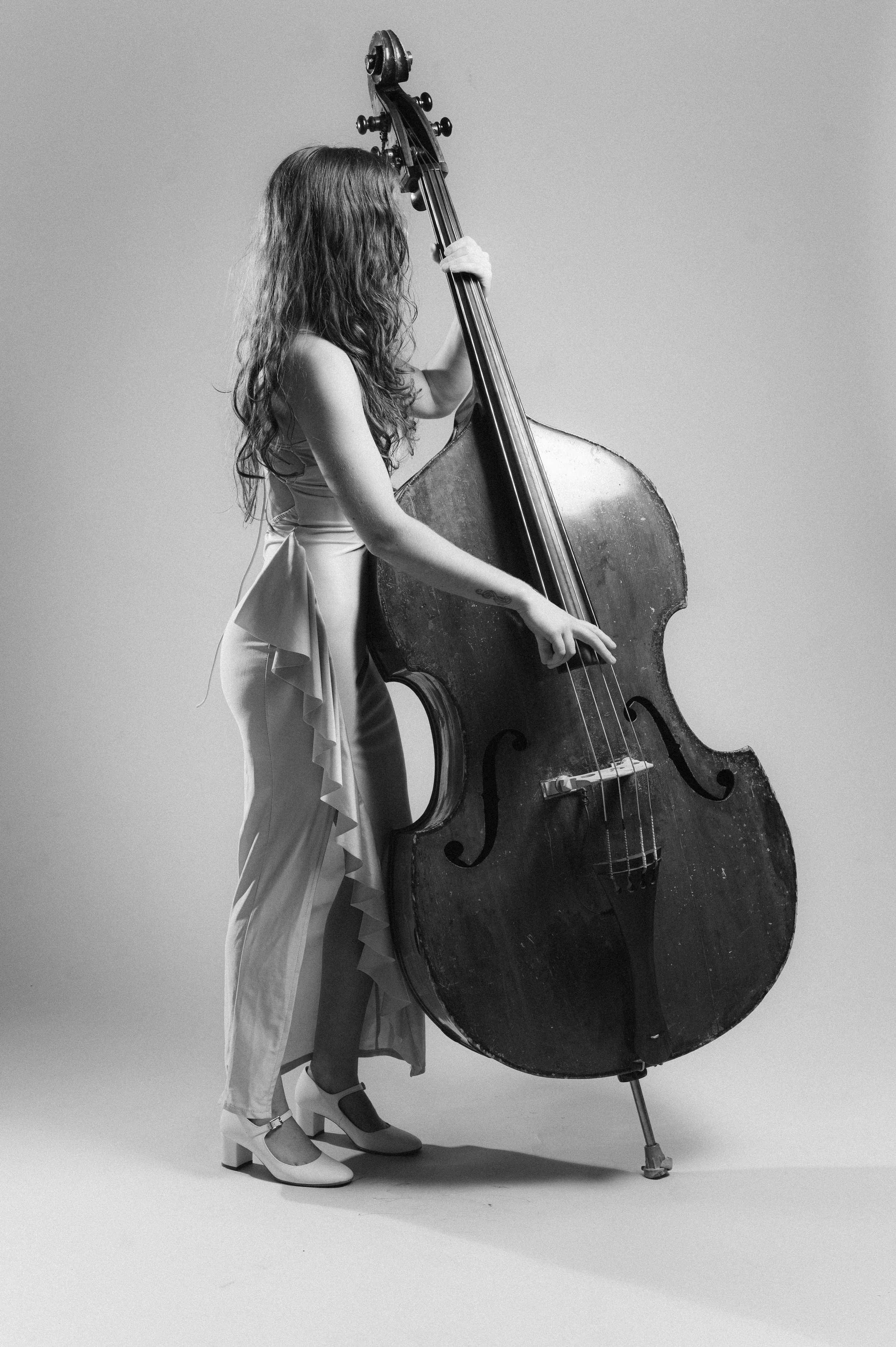 Black and white photo of a woman with long curly hair wearing a sleeveless blouse, wide-legged pants, and heels, standing next to a large double bass.