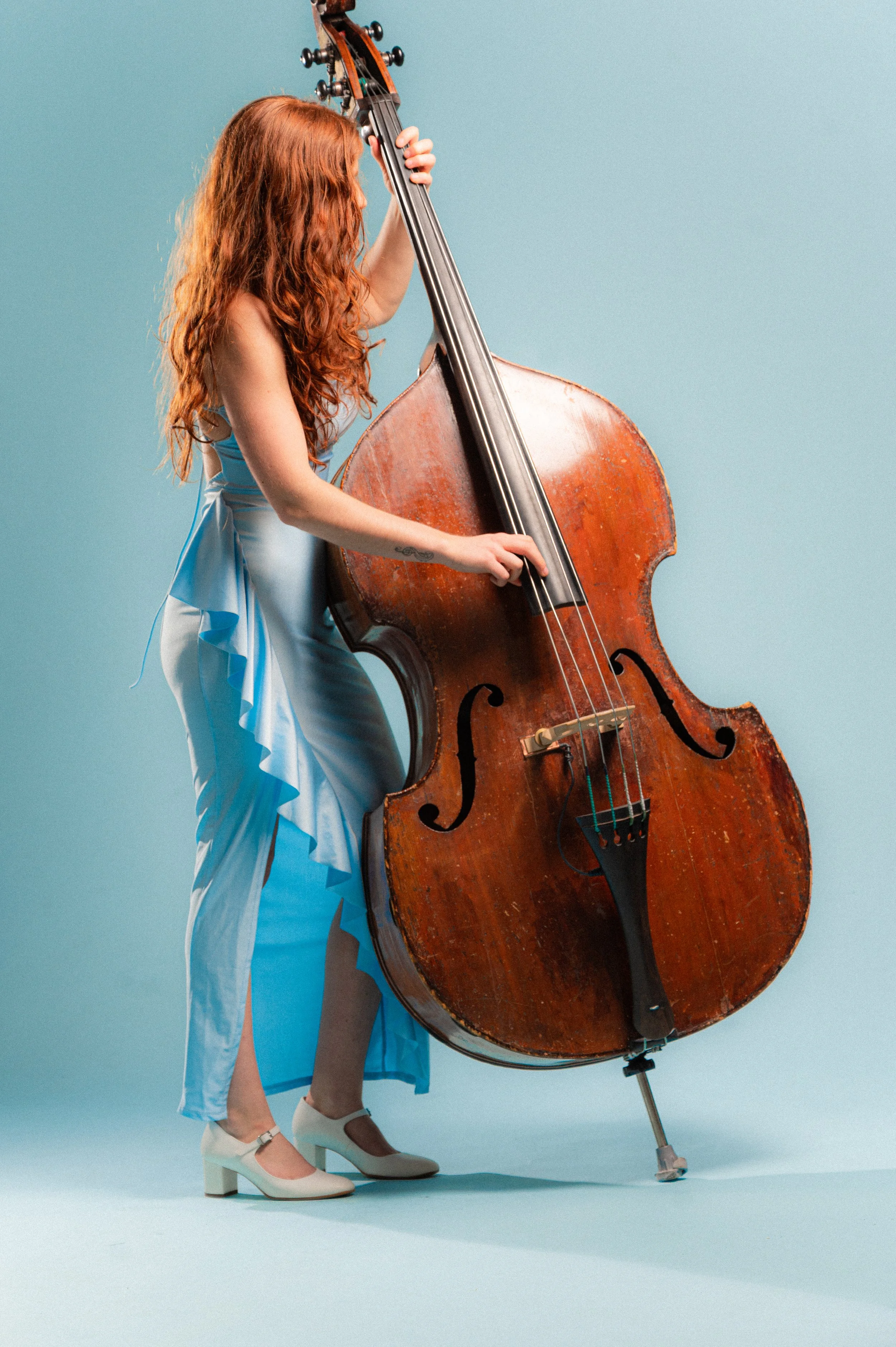A woman with long red curly hair playing a large upright double bass against a light blue background. She is dressed in a blue dress with ruffled details and wearing white shoes with a block heel.