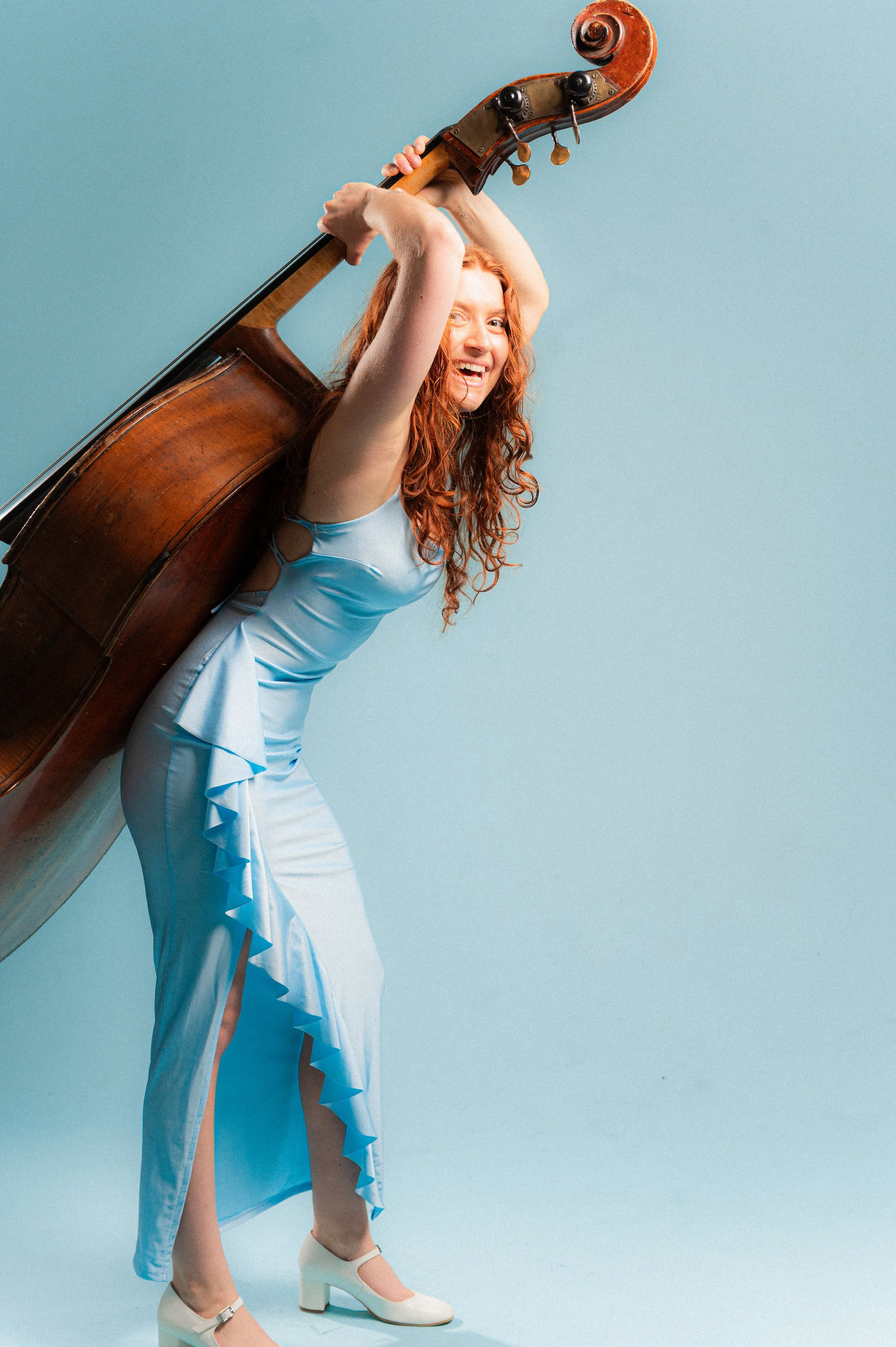 A woman with long red curly hair wearing a light blue dress with a high slit, white high heels, and holding a large vintage wooden cello over her shoulder, smiling at the camera, against a solid baby blue background.
