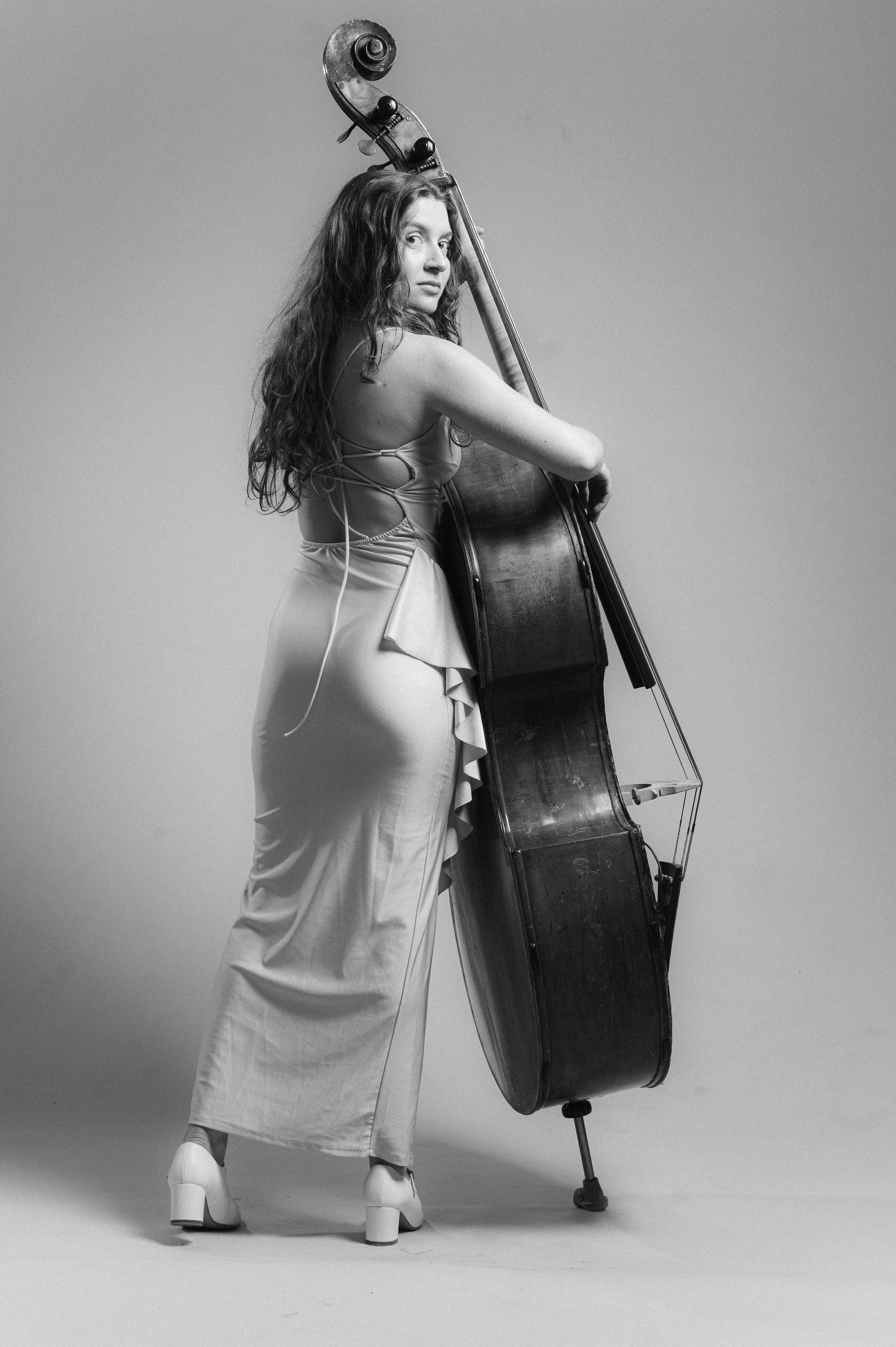 Black and white photo of a woman with long wavy hair standing sideways, holding a large upright double bass. She is wearing a flowing dress with open sides and high heels with chunky heels. The background is plain and uncluttered.