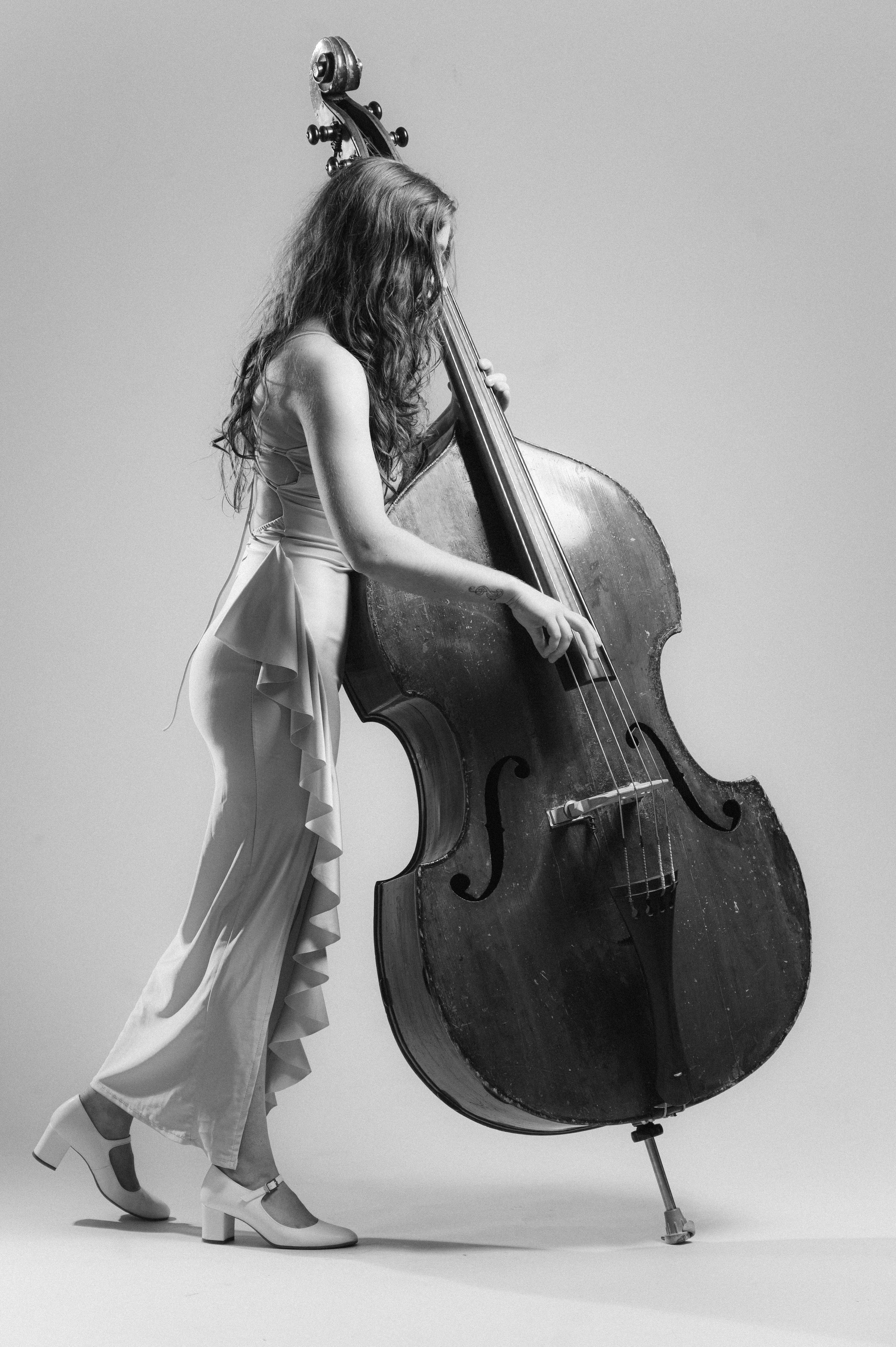 Black and white photo of a woman with long curly hair playing a large upright double bass against a plain background. She is dressed in a light-colored dress with ruffled details and wearing high-heeled shoes.