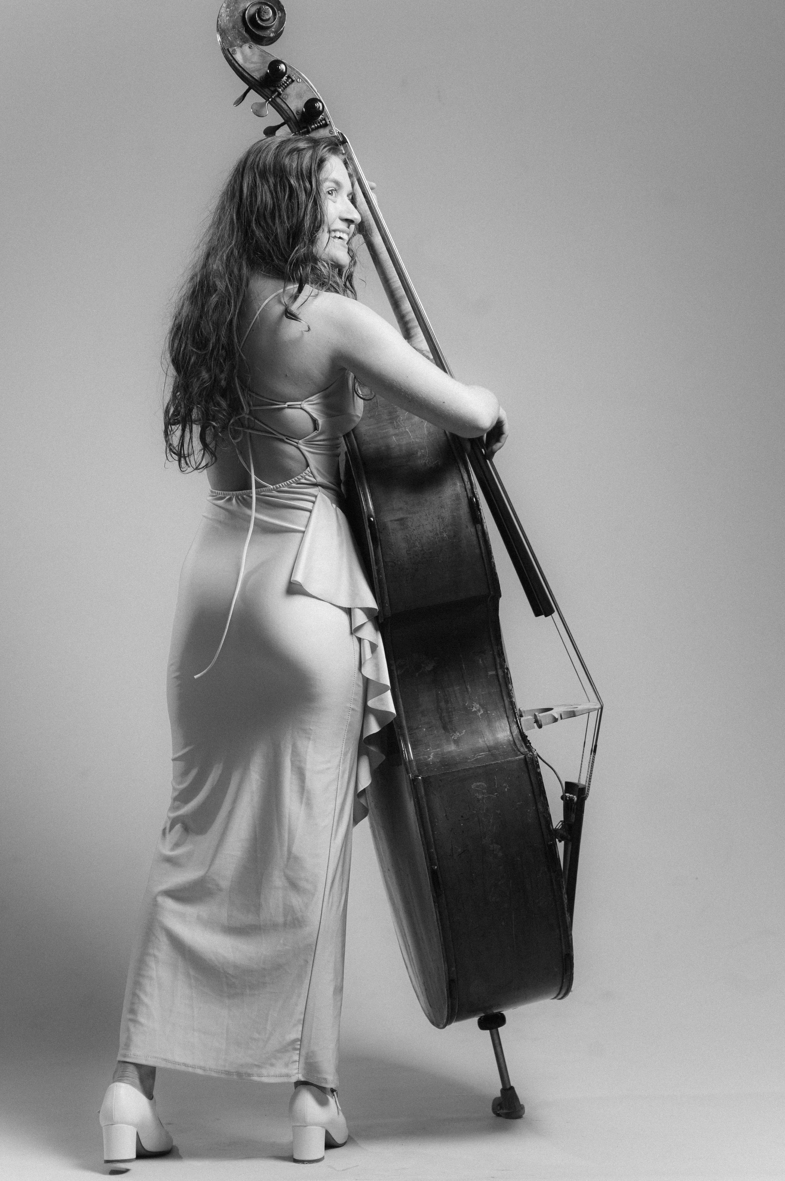 A woman with long curly hair in a dress and high heels poses with a large upright bass, smiling and looking to her right, against a plain background.