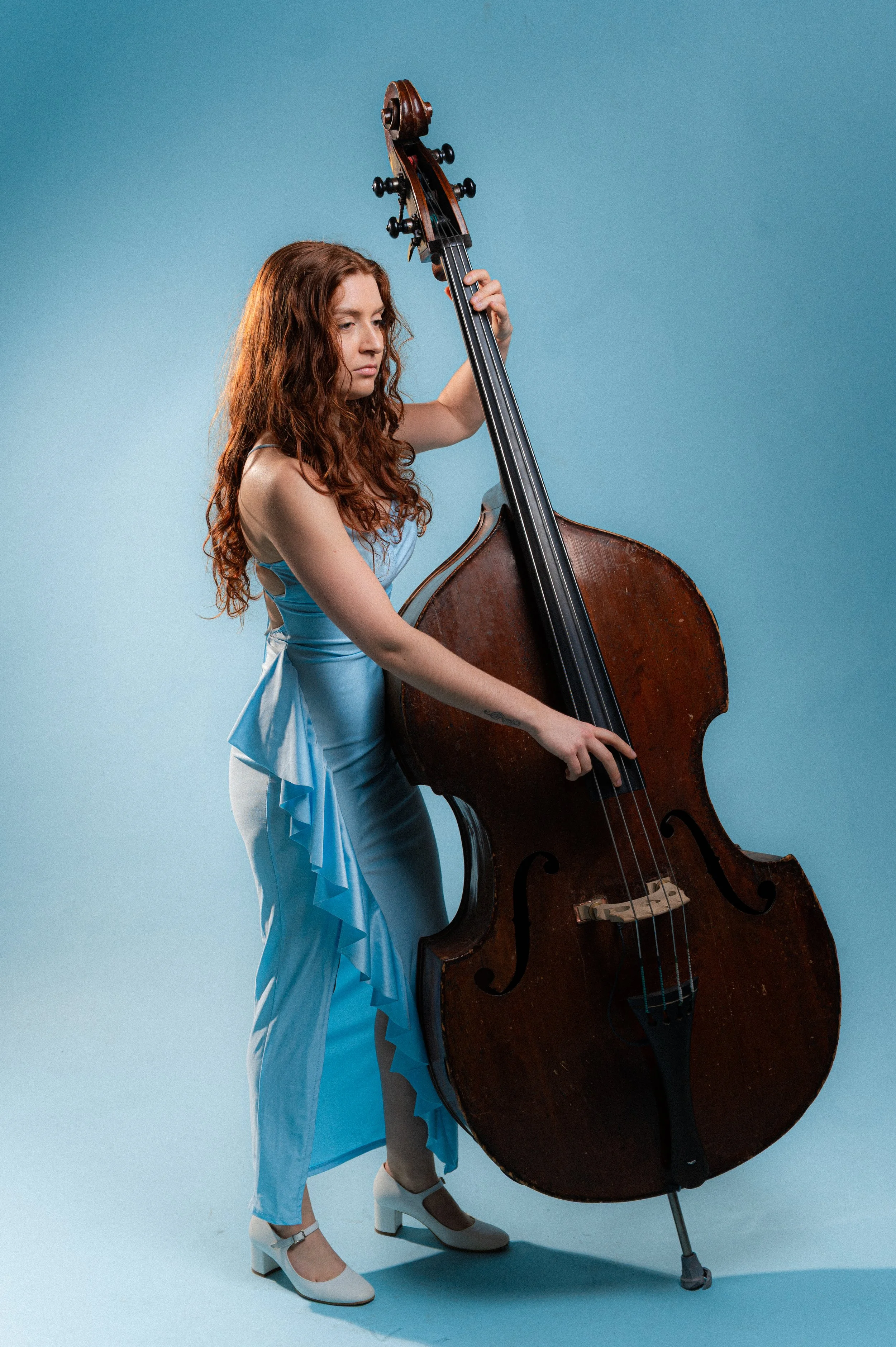 A woman with long curly red hair wearing a light blue dress with ruffles and white high heel shoes is playing a large, vintage wooden double bass against a light blue background.