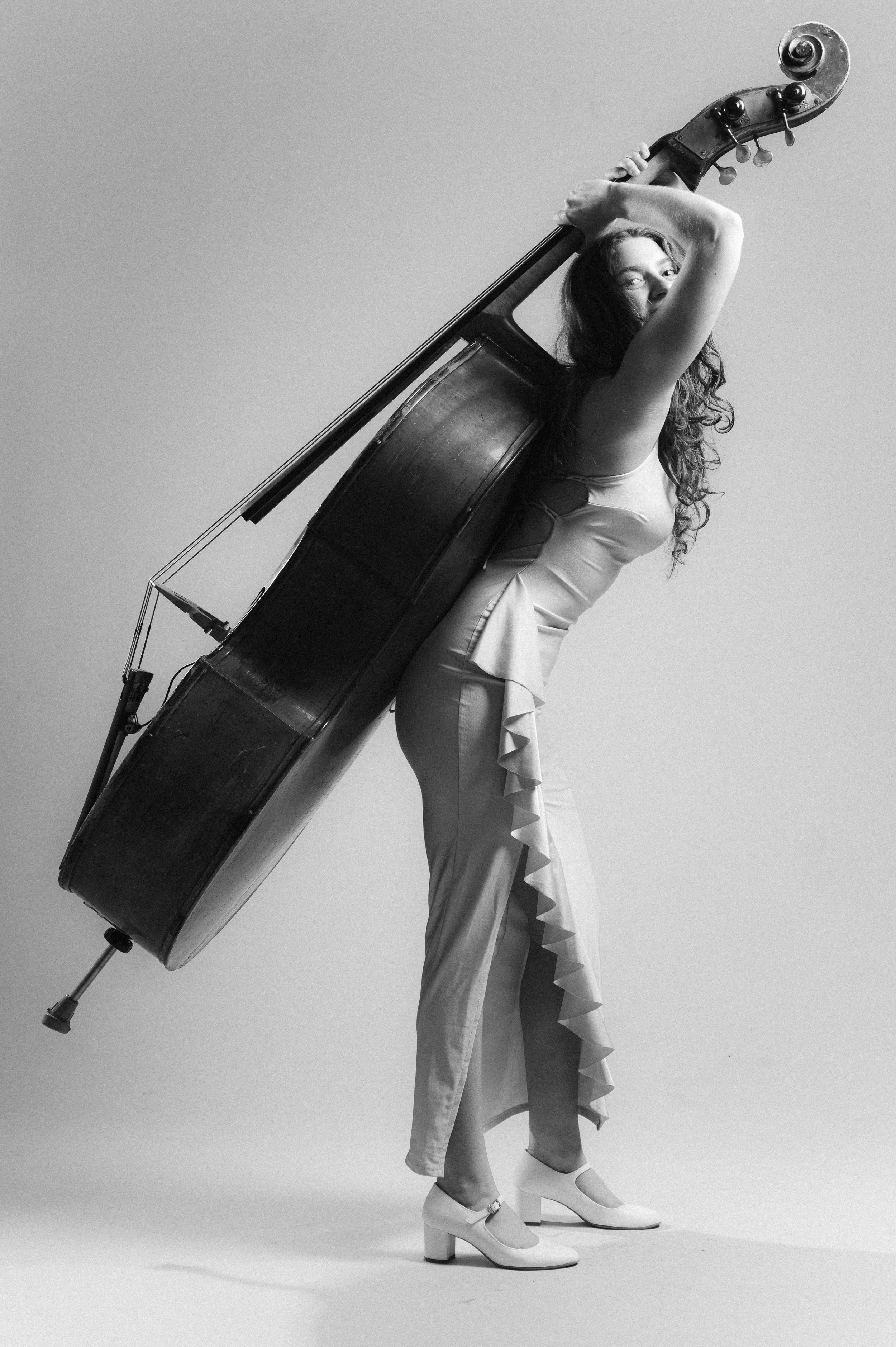 Black-and-white photo of a woman holding a large upright double bass, with her arms around it. She is wearing a satin dress with ruffles and high-heeled shoes. The woman is looking at the camera with a slight smile, against a plain light background.