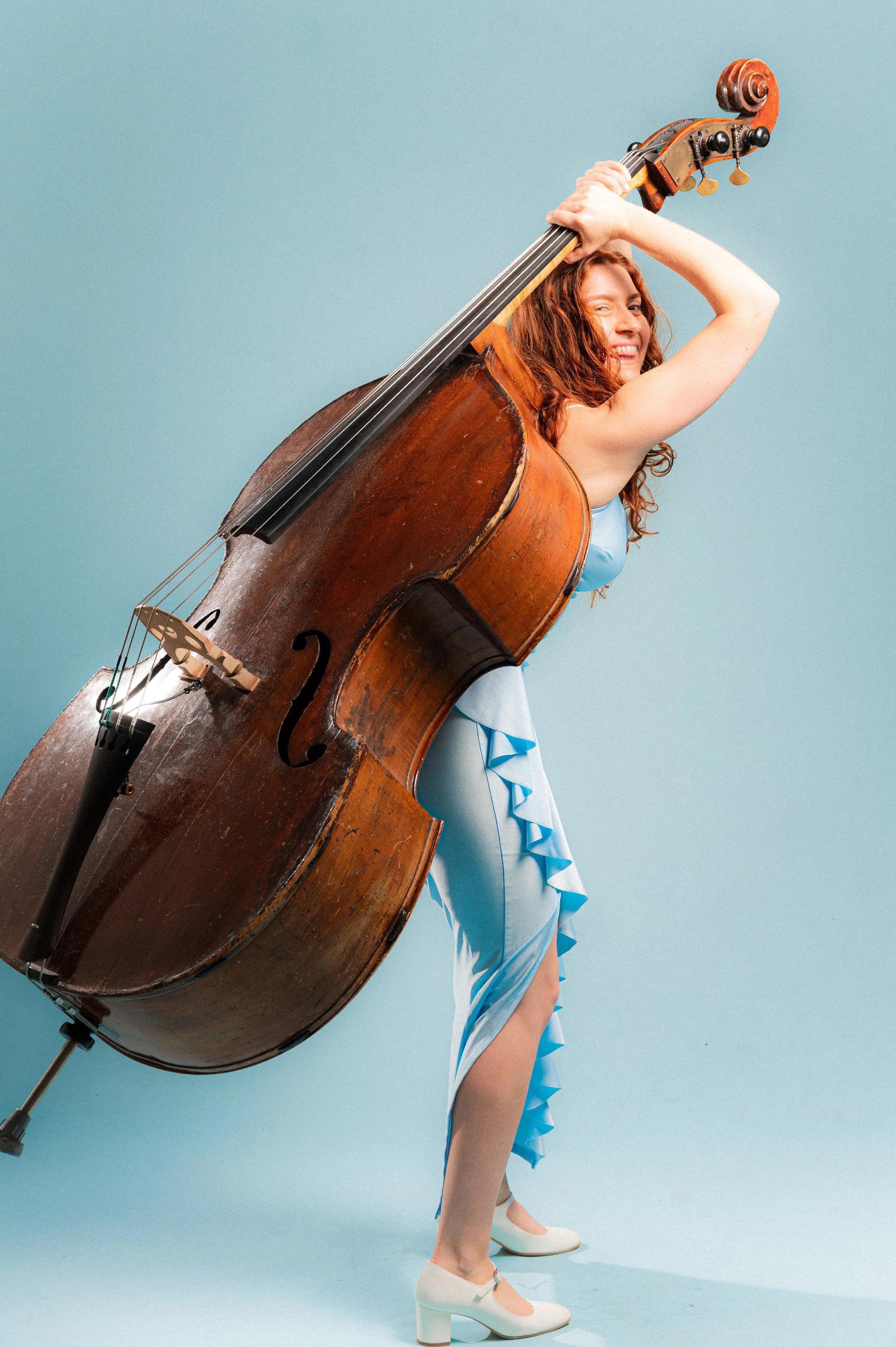 A woman in a light blue dress with ruffles, wearing white heeled shoes, poses against a light blue background while holding a large vintage-looking double bass.