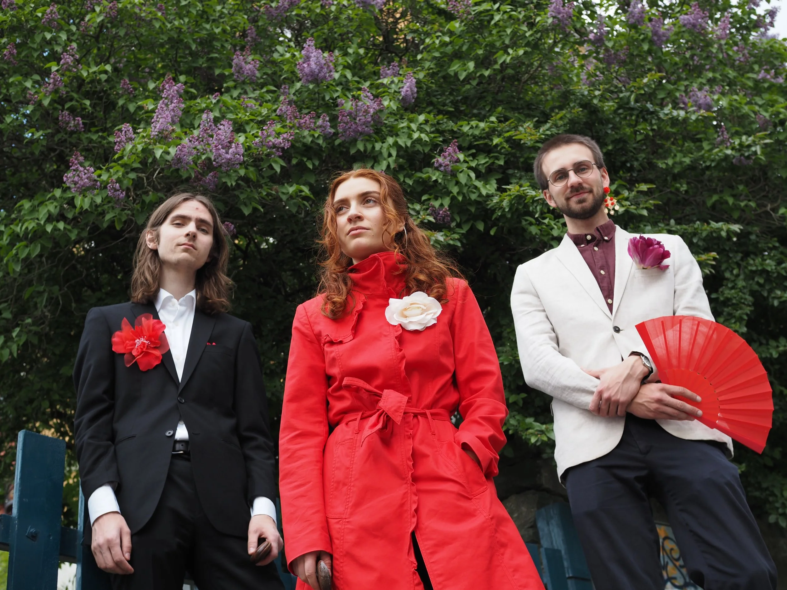 Three people standing outdoors in front of green foliage with purple flowers. The person on the left is a man with long brown hair wearing a black suit with a white shirt and a red flower pinned to his jacket. The middle person is a woman with red hair wearing a red coat with white flowers on the chest. The person on the right is a man with glasses and a beard, wearing a white suit with a purple shirt, red earrings, and holding a red fan.