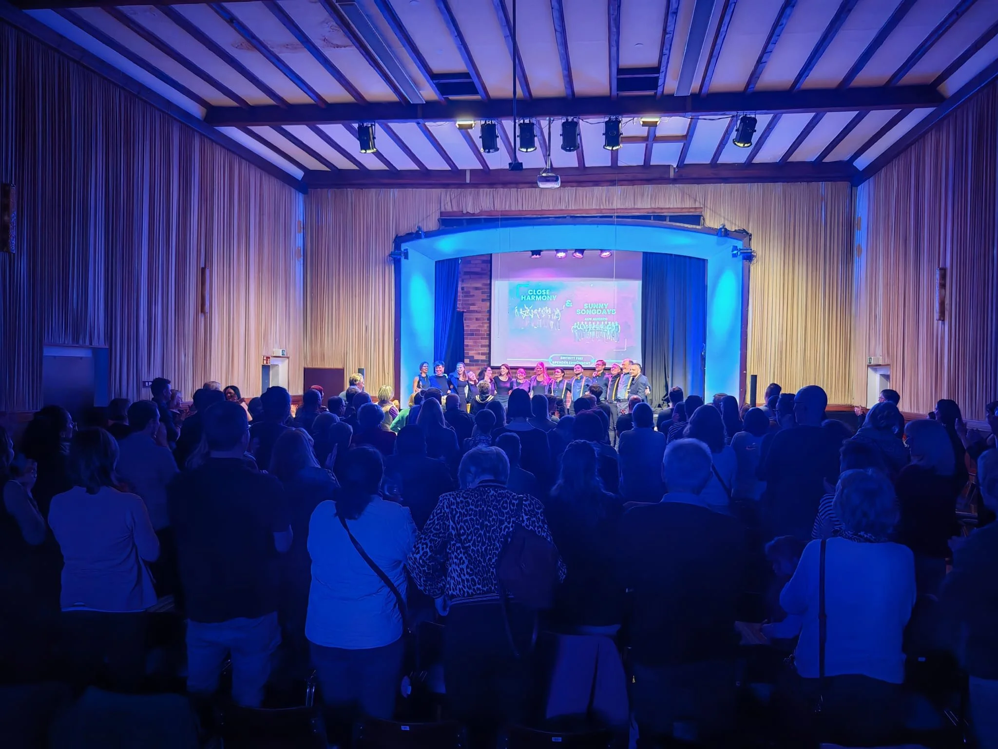 Ein Saal mit einem Publikum, das einem Chor beim Singen zuschaut. Im Hintergrund ist eine große Leinwand mit Text und bunten Lichtern. Der Raum ist mit Holz verkleidet, die Decke ist mit Holzbalken gestaltet, und es gibt blaue Beleuchtung.