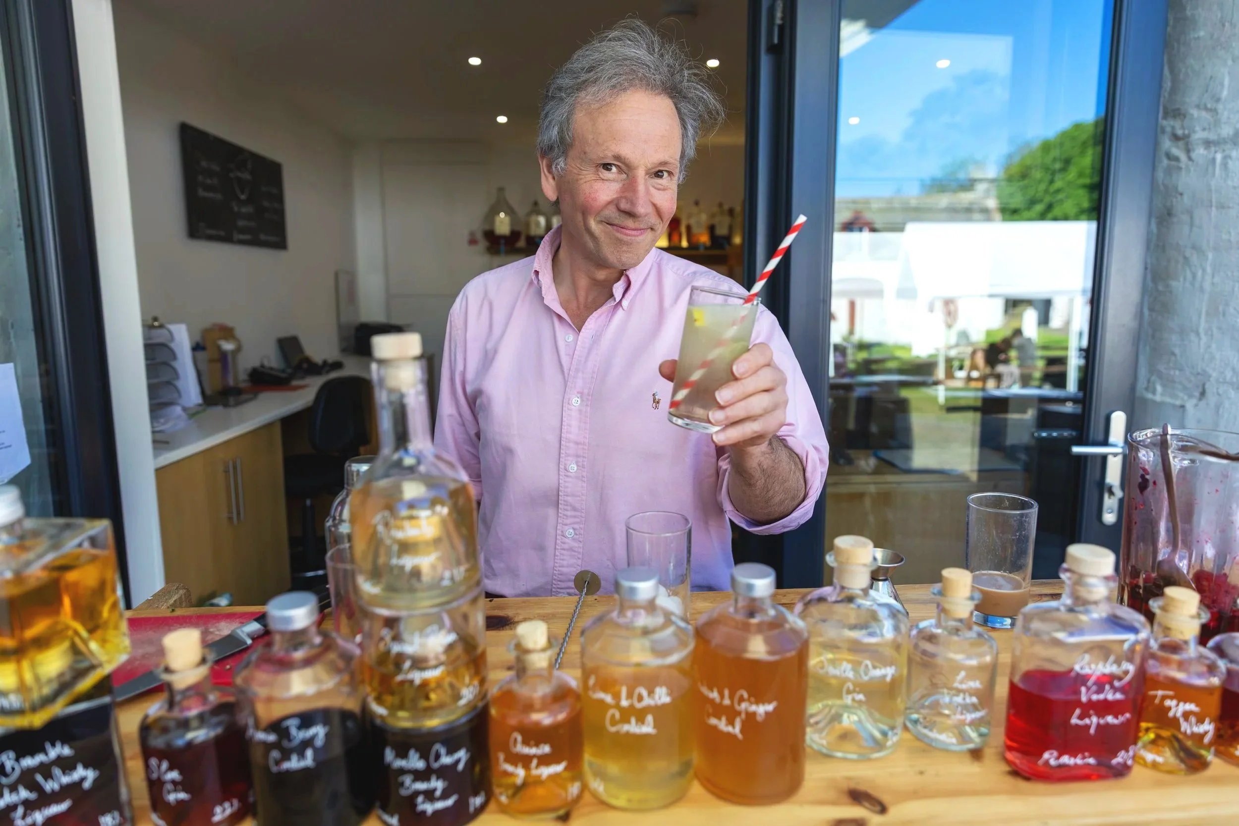 A middle-aged man with gray hair, wearing a pink button-up shirt, smiling and holding a glass with a striped straw, standing behind a display of various flavored liquids in glass bottles, inside a brightly lit room with large windows.