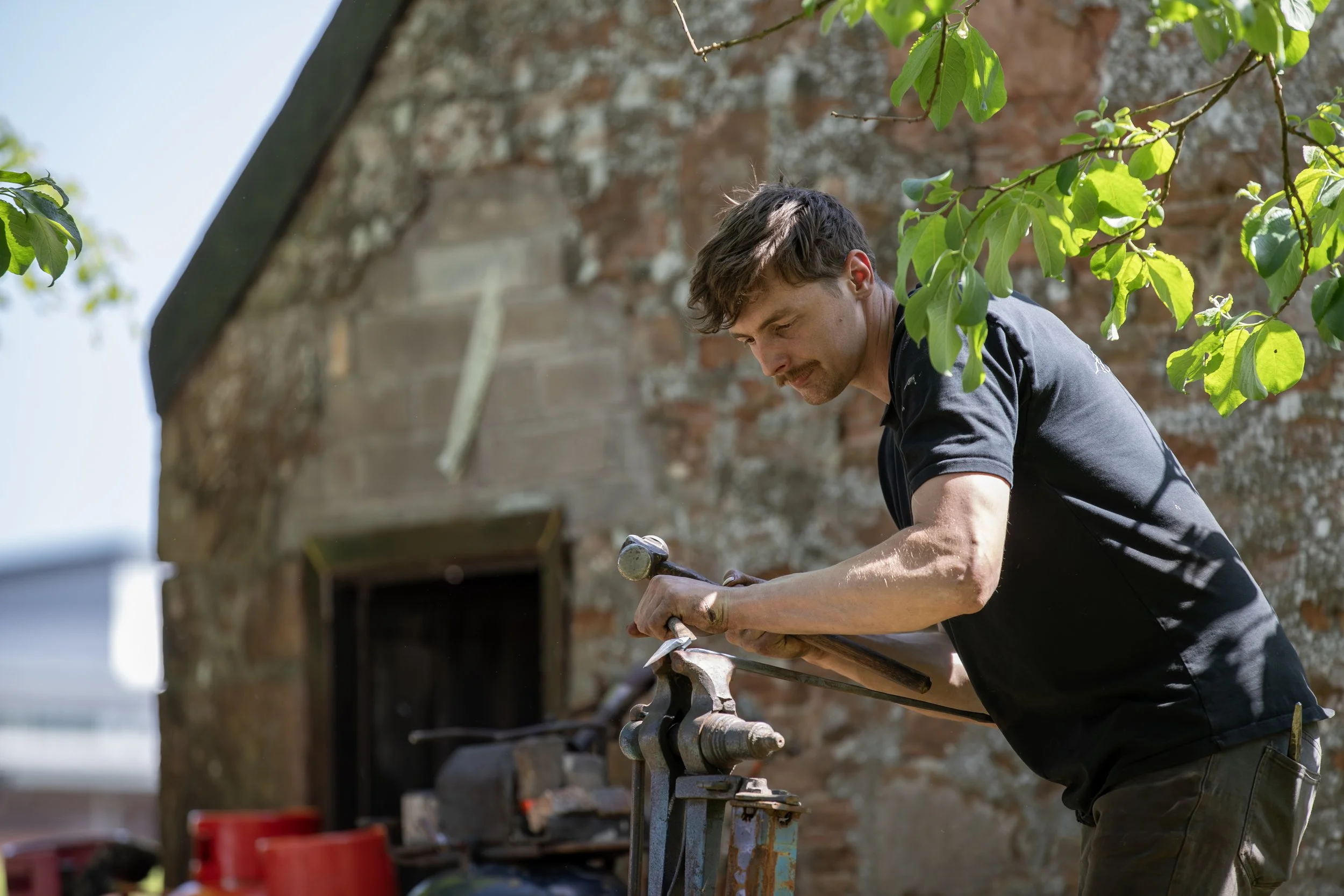 A man working outdoors on a metal project, using a hammer and chisel, with a brick wall and green leaves in the background.