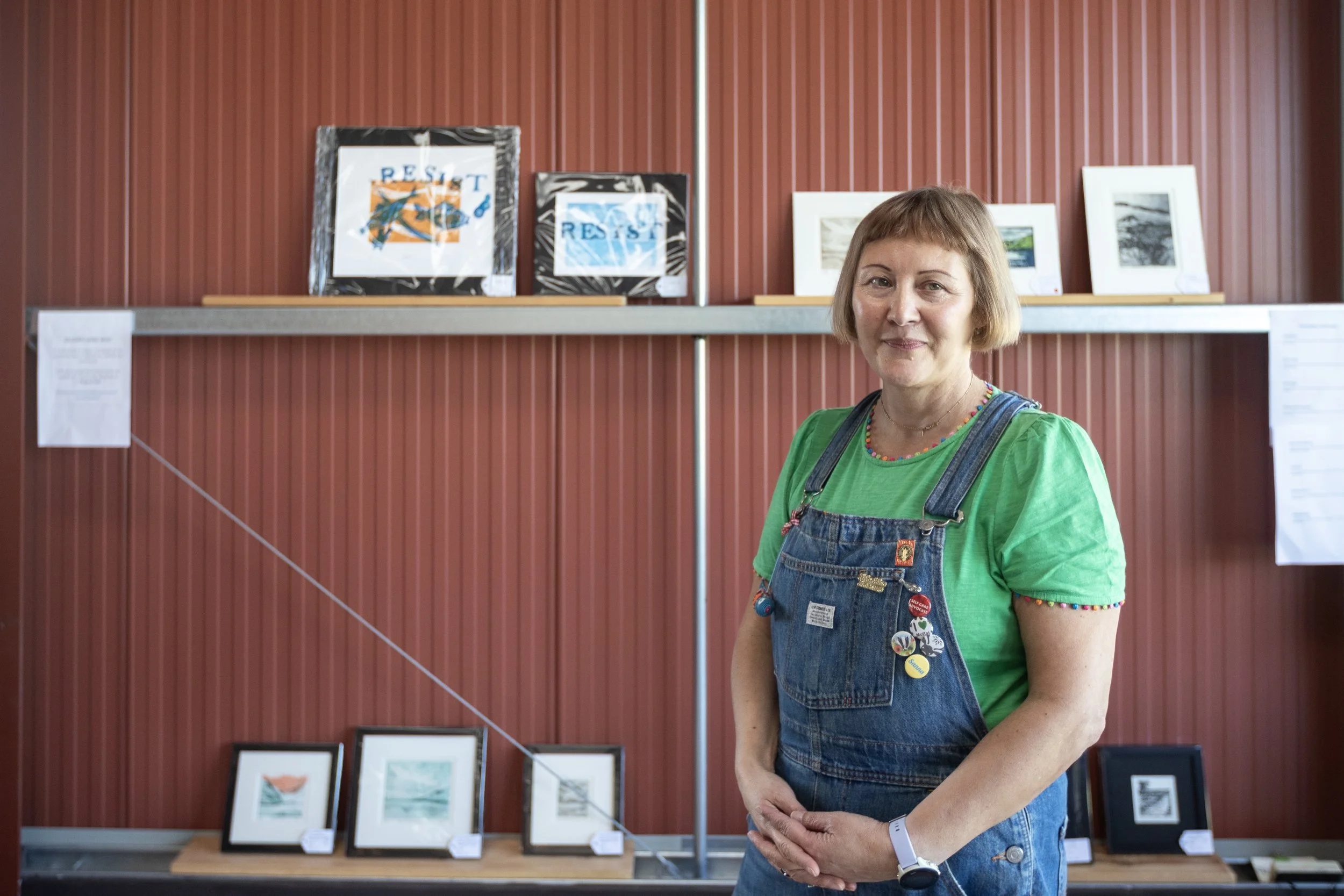 A woman with short light brown hair smiling at an art exhibition, wearing a bright green t-shirt and denim overalls with buttons and badges, standing in front of a display of framed artwork against a reddish-brown wall.