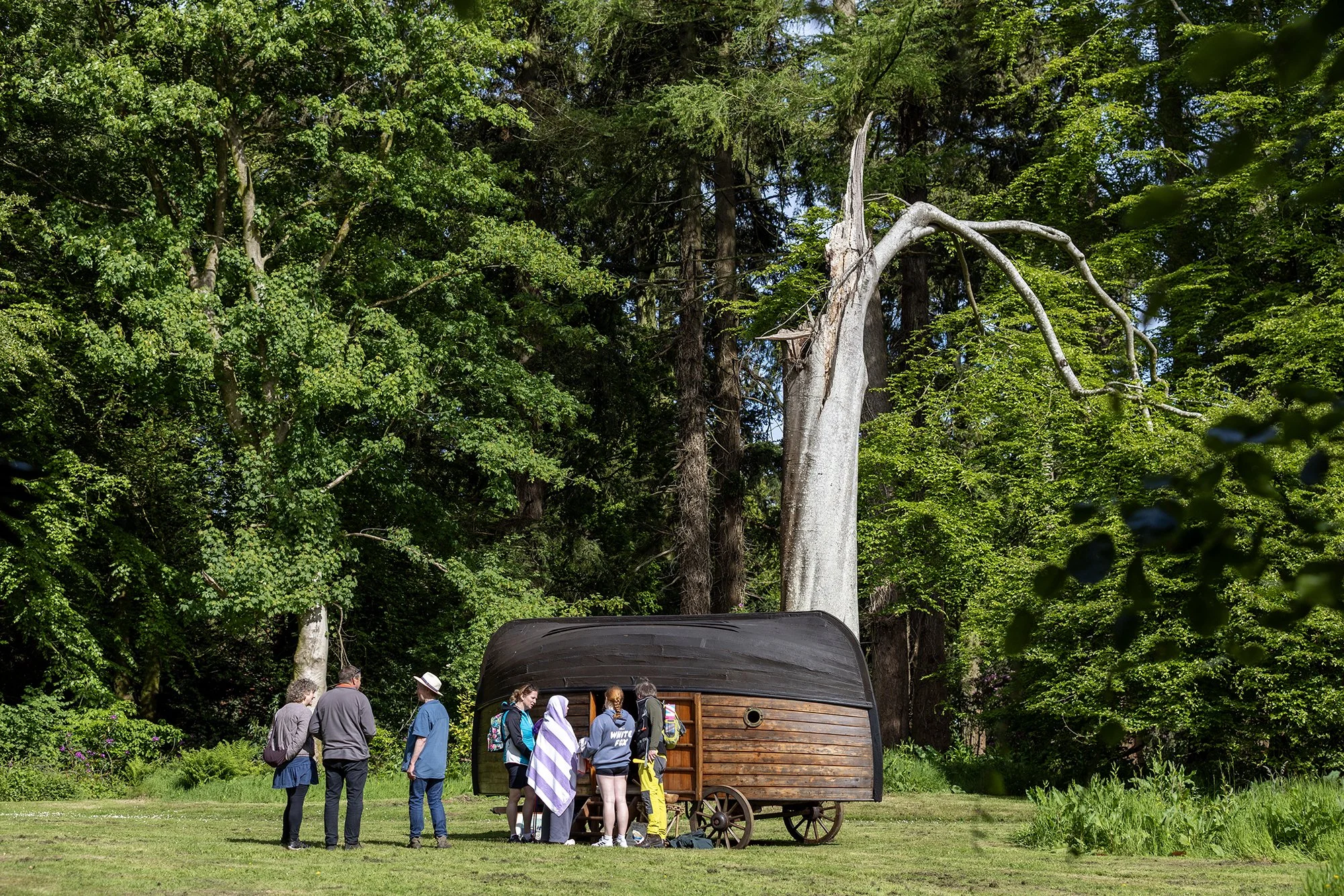 A group of people standing outdoors near a small wooden wagon with a curved roof, set in a lush green forest with tall trees and bright foliage.